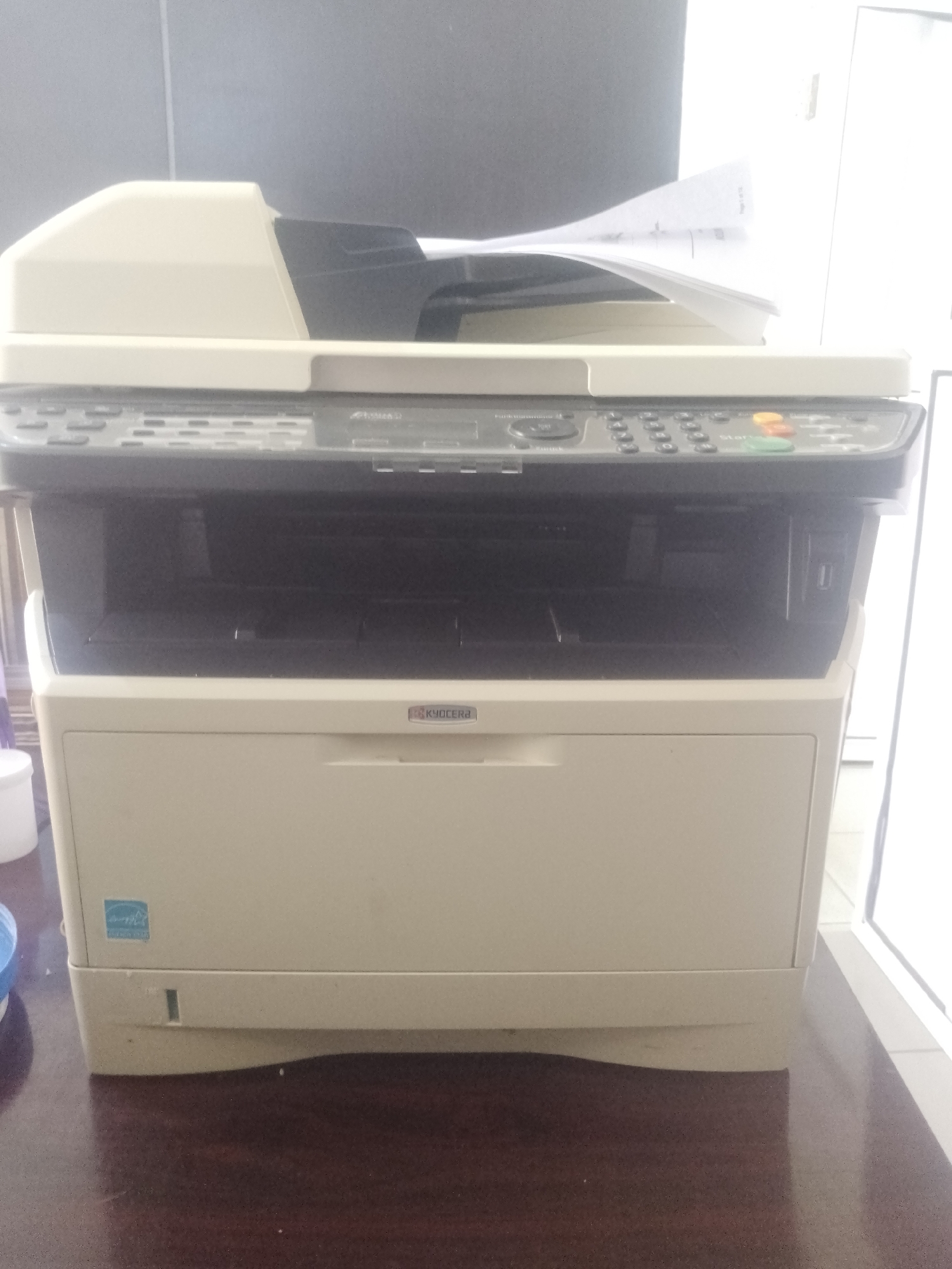 An office printer is centrally located on a dark wood desk. The printer is a Kyocera brand, beige and black in color, with a document feeder on top. The printer has a control panel with buttons and a small display screen. A stack of papers is visible in the document feeder. The background is a gray wall with a window on the right side, allowing bright light to enter the room. The overall environment appears to be an office in Gura Top, Nigeria.