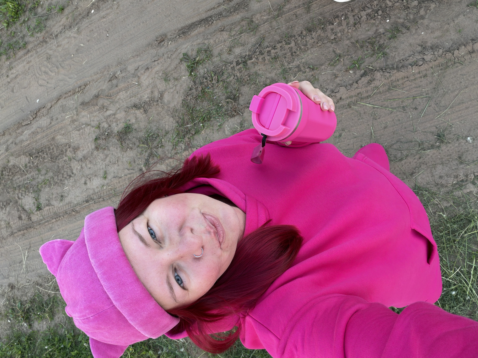 An overhead selfie, taken from an upward angle, captures a young adult looking directly at the camera with a slight smile. The subject has reddish-brown hair, blue eyes, and fair skin, and wears a silver hoop nose ring on their left nostril. They are dressed in a vibrant pink hooded sweatshirt, which appears to have cat-ear details on the hood, and a matching pink beanie or hat underneath.
In their right hand, they hold a bright pink thermal mug with a black strap from which a small, brown, whistle-like charm is suspended. The person's fingernails are painted with a subtle, light-colored polish.
The background is an outdoor setting featuring a light brown, unpaved dirt path or road, marked with tire tracks and interspersed with patches of green grass and dry plant matter. The lighting suggests it is daytime, with no harsh shadows indicating a bright or overcast sky. The overall mood conveyed by the subject's expression is relaxed and pleasant.
No visible text is present in the image.