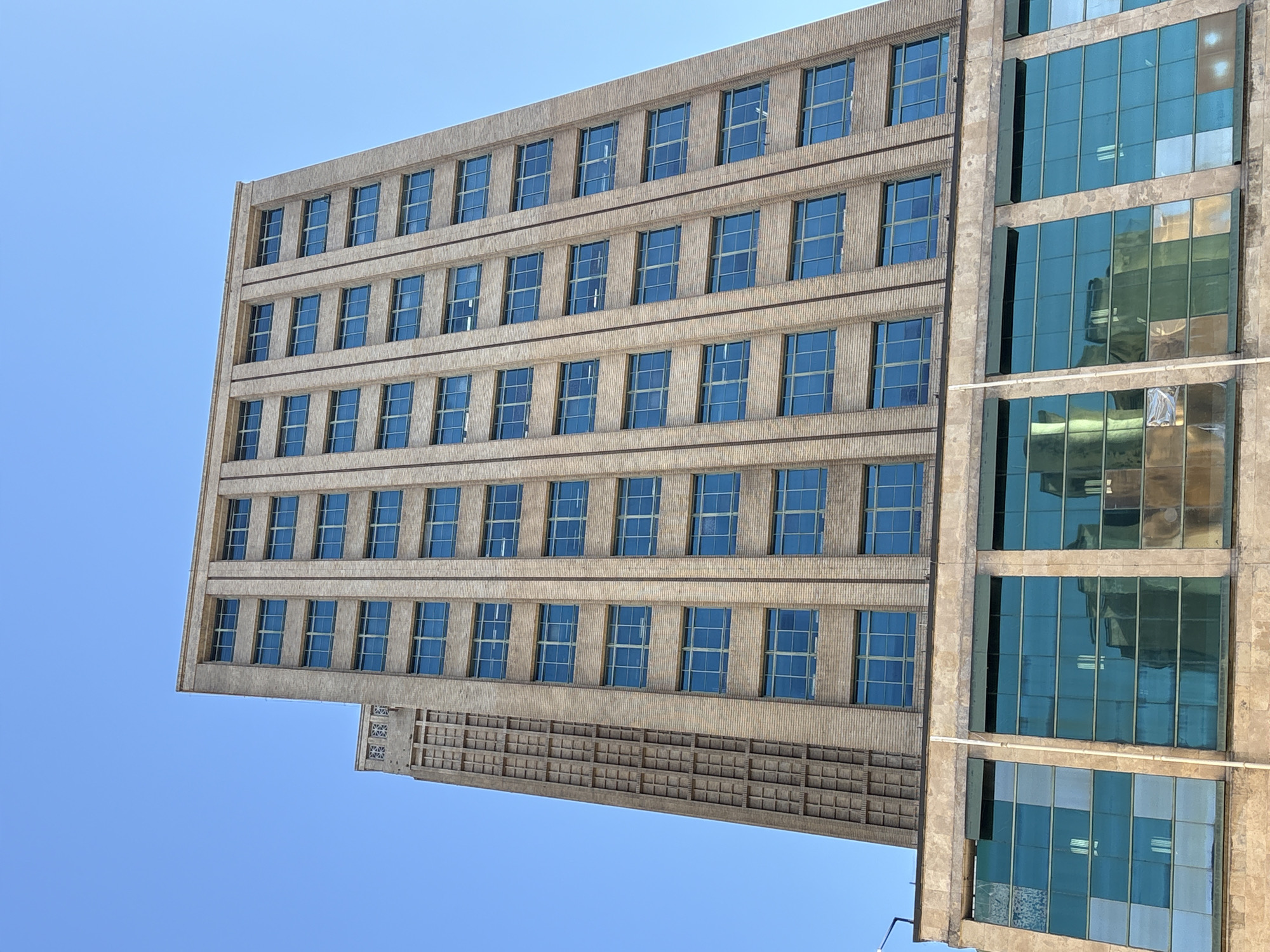 The image shows the exterior of a tall, modern office building, with a clear blue sky as the backdrop. The building is constructed with light-colored brick and features numerous large, rectangular windows with teal-colored frames, arranged in a grid pattern across its facade. The lower section of the building has a different architectural style, with larger, more expansive windows that reflect the surrounding environment, suggesting a commercial or public space. The building appears to be located in an urban setting, as implied by its scale and design. The specific location in São Paulo, Brazil, is not visually evident from the image alone, but the architectural style is consistent with contemporary urban development. No people are visible in the image.
