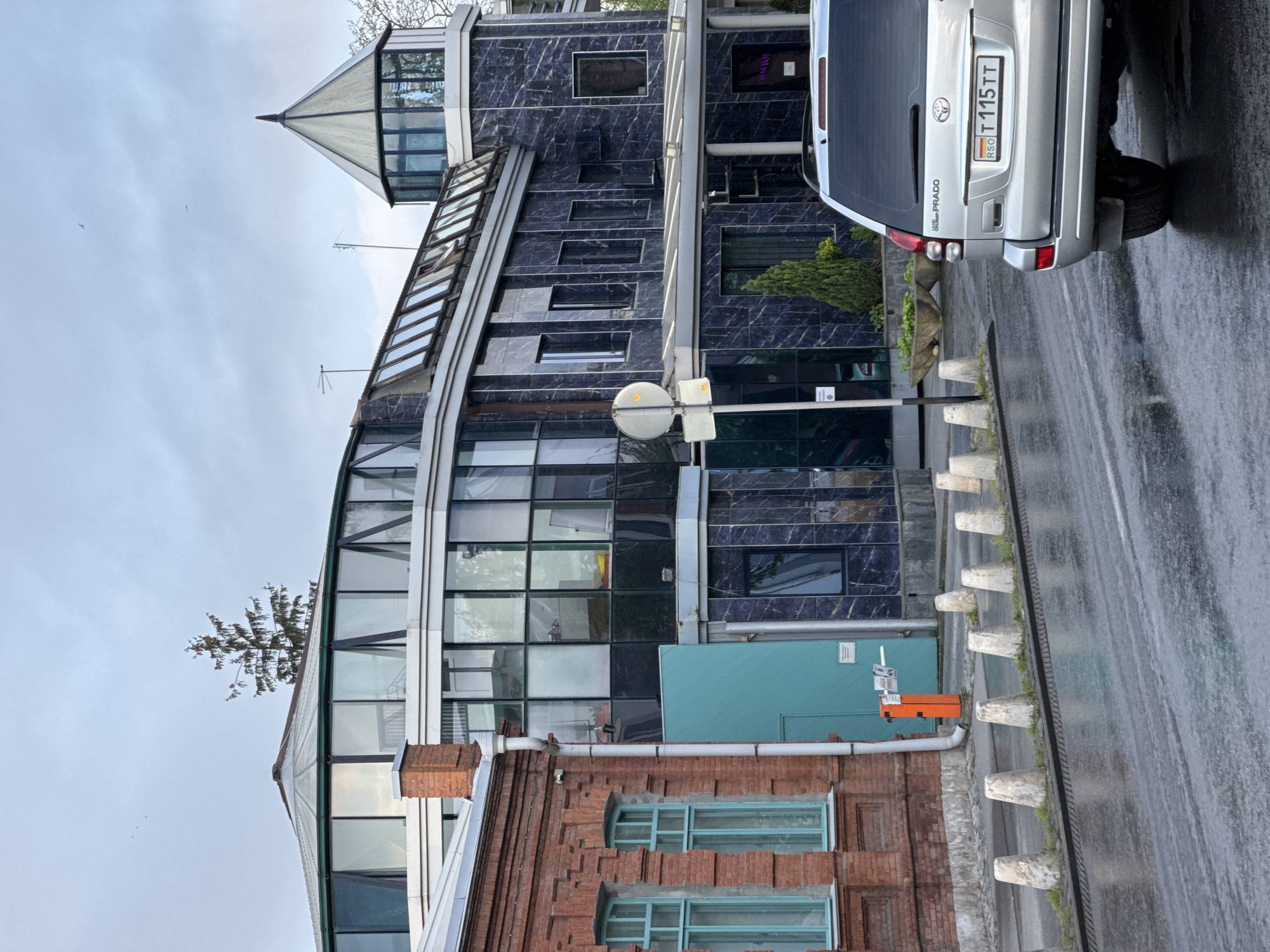 The image shows a multi-story building complex in Akhieli, Georgia. The architecture is a mix of styles, with an older brick facade featuring light blue-framed windows at the base, transitioning into a modern glass and steel structure. Above this is a section with dark blue tiled walls with rectangular windows, topped by a glass-enclosed turret.
To the right of the building is a silver Toyota Land Cruiser Prado parked on the side of the street. The license plate is visible: RSO T 115 TT. The road surface is wet, suggesting it has recently rained. The sky is overcast. Concrete bollards line the edge of the sidewalk next to the road. A street sign is visible. An orange box and a blue utility box are also present near the brick building. A small tree is growing on top of the corner between the glass section and the brick building.