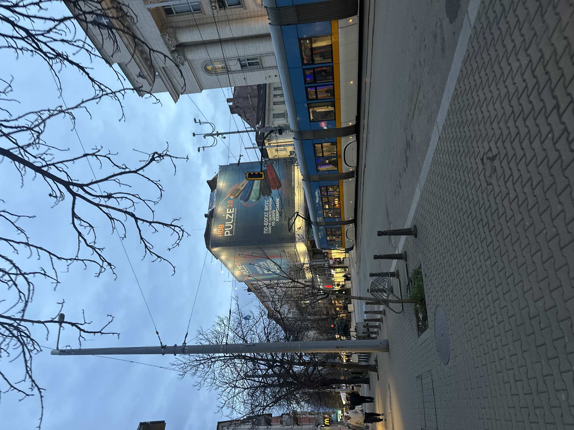 The image captures an urban street scene in Sofia, Bulgaria. A blue and white tram is prominently featured running along the street. A large billboard advertising "PULZE 3.0" is visible on a nearby building. The sidewalk is paved with grey interlocking bricks. Bare trees line the street. Street lights are present along the sidewalk. Several people are walking on the sidewalk.