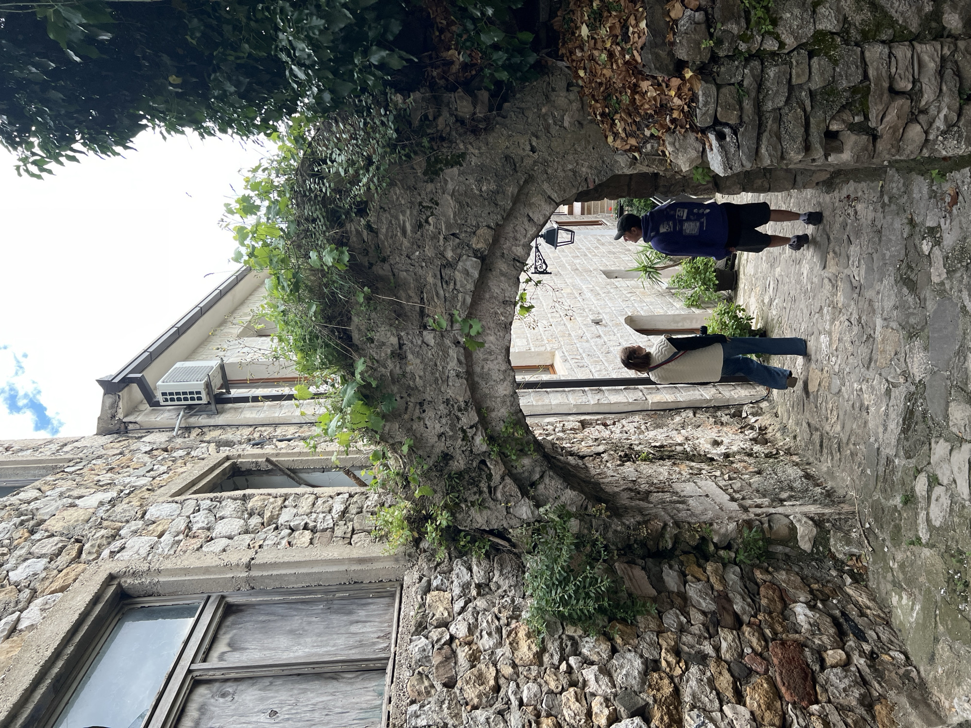 The image captures a stone archway in Ulcinj - Ulqin, Montenegro. The arch is partially covered with vegetation, situated between two buildings constructed of stone. Through the archway, two people are visible: one wearing a blue jacket, black shorts and a baseball cap, walking away from the camera, and another with a light-colored top and blue jeans, seemingly using a phone or looking down. The buildings flanking the arch are also made of stone, and one has broken windows and an air conditioning unit. The ground under the arch is paved with stone.