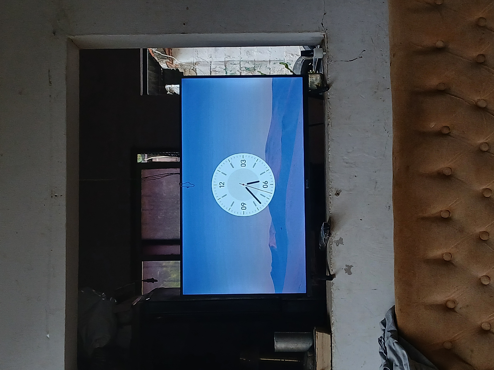 The image shows a television screen displaying a clock face with a minimalist landscape background. The clock has hour markers and hands, with the time appearing to be approximately 5:30. The television is situated within an opening in a rough, unfinished wall, possibly a window or a doorway. Below the television, the top of a tufted, orange couch is visible. The surrounding environment appears to be indoors, with exposed concrete walls and some debris. The lighting is dim, suggesting it is daytime but the area is not well-lit. The location is identified as Tacarigua, Venezuela.