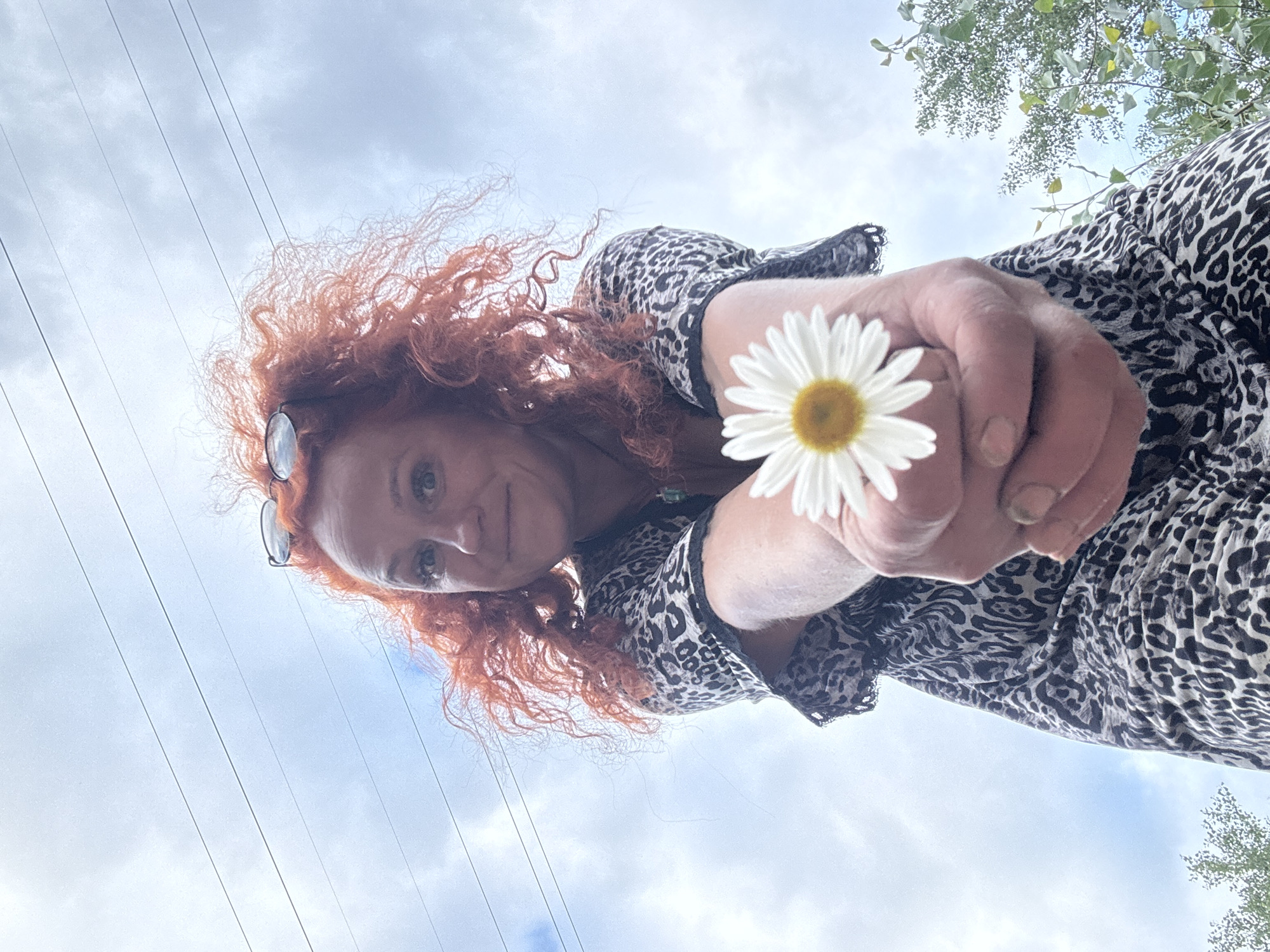 A woman with long, curly red hair, wearing a leopard print dress, holds a daisy up to the camera, her face angled towards the sky. She wears round-framed glasses perched on her head. The image is taken from a low angle, looking up at the woman against a backdrop of a cloudy sky with visible power lines stretching across the frame. The woman's expression is direct and engaging, with a slight smile. The setting appears to be outdoors, with some green foliage visible at the top right, suggesting a natural or park-like environment. The overall mood is bright and cheerful, with the daisy adding a touch of nature and simplicity to the composition. The time of day seems to be daytime, given the natural light and cloudy sky. There is no visible text in the image.