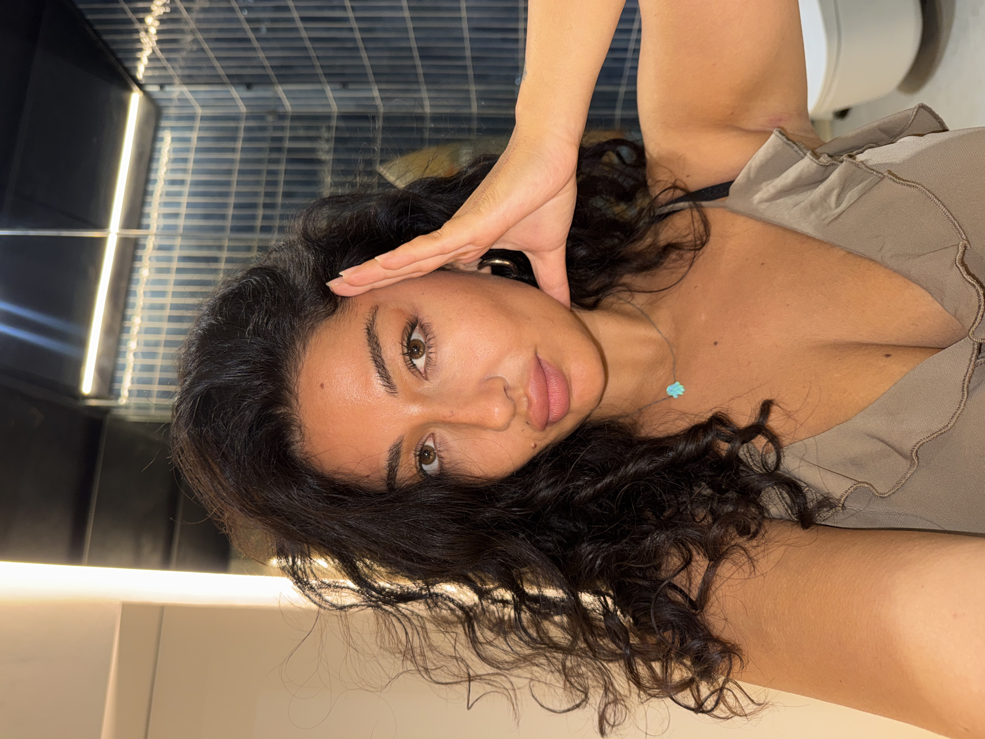 An upward-angled, close-up selfie captures a young woman with dark, curly hair and an olive skin tone, looking directly at the camera with brown eyes and a neutral expression. Her right hand is raised, with her fingers touching her temple area. She wears a light brown or taupe top with ruffled details and a visible black bra strap. A delicate necklace with a small, light blue pendant, possibly turquoise and shaped like a flower or cross, adorns her neck. Notable facial features include moles on her upper lip and near her chin.
The background suggests an indoor setting, likely a modern bathroom or hallway, characterized by artificial lighting. Reflective surfaces with linear patterns, possibly tiles or grilles, are visible. Strips of fluorescent lighting, both vertical and horizontal, illuminate the space. One crop reveals a portion of a white fixture, which appears to be a toilet, further indicating a restroom environment. The overall ambiance is contemporary and well-lit. There is no visible text in the image. The location context is Kuta Selatan, Indonesia.