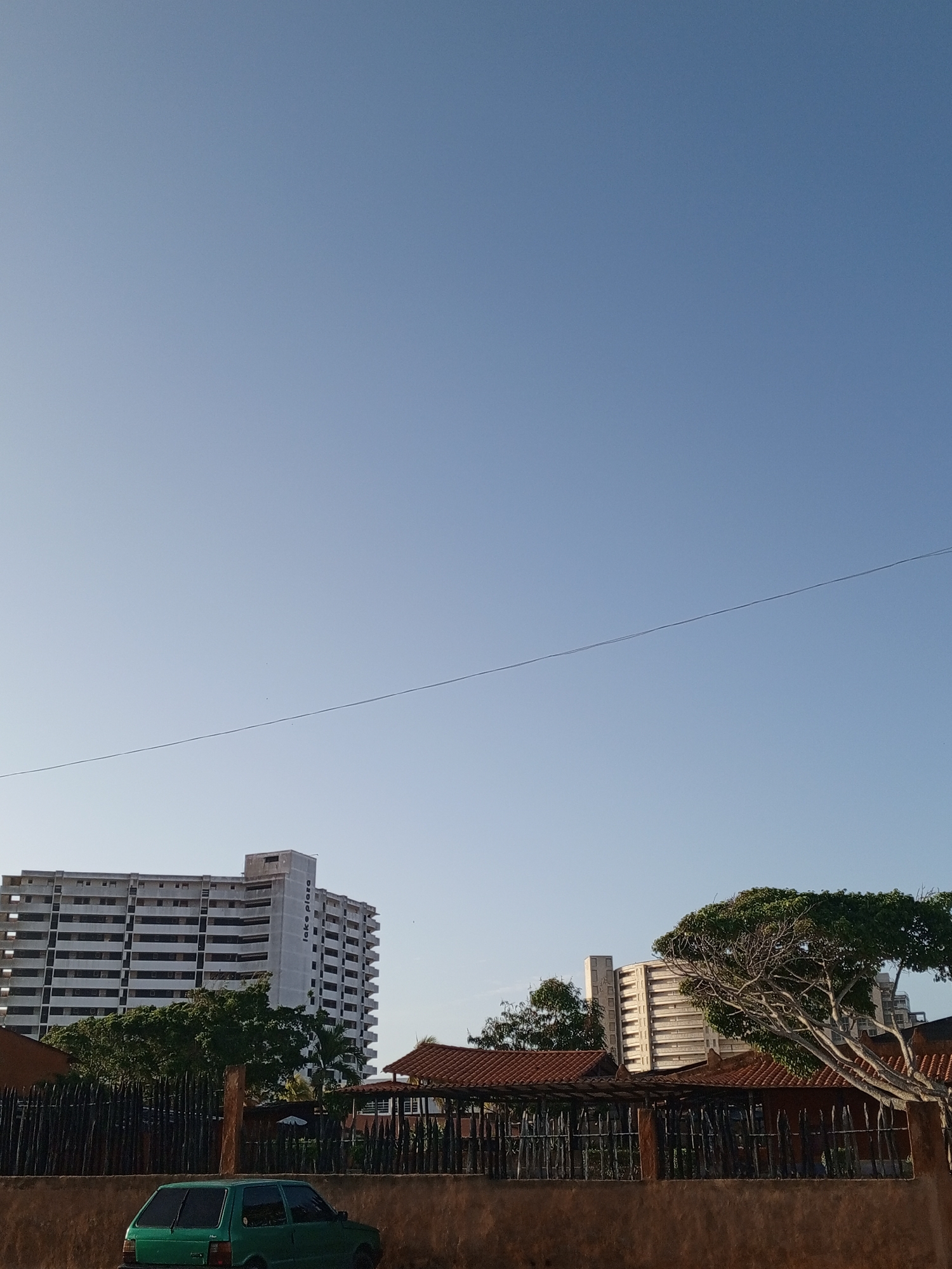 The image captures a street scene in Porlamar, Venezuela. In the foreground is a low wall, behind which stands a dark wooden fence enclosing a building with a red tile roof. A small, green car is parked near the wall.
Behind the fence and building, several multi-story buildings dominate the skyline. One building, identified by the sign as "Lake Plaza," is taller than the others. The sky is a clear, light blue, with a power line stretching across the view. The lighting suggests early morning or late afternoon. No people are visible in the frame.