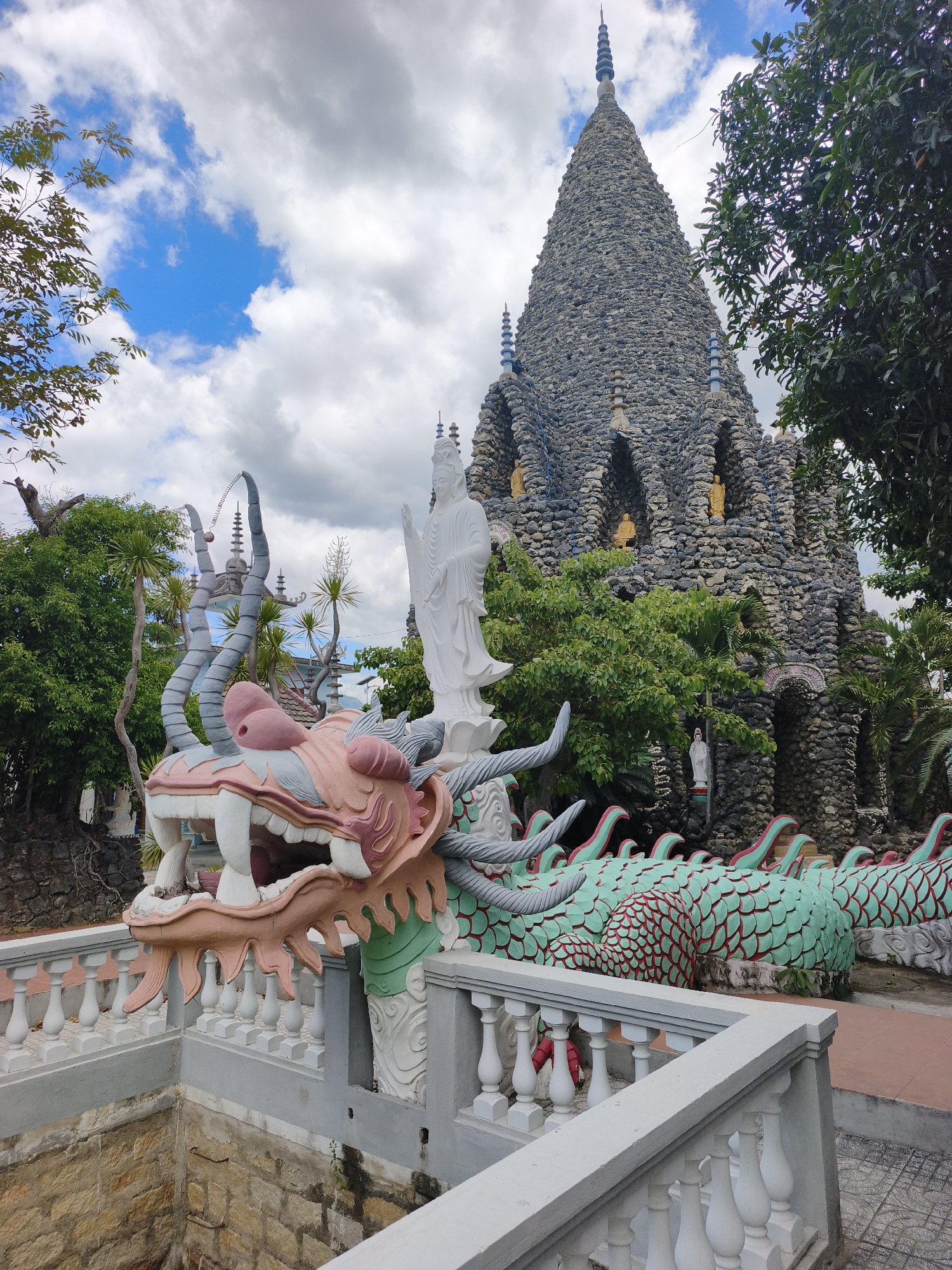 The image shows a large, elaborate stone structure resembling a temple or pagoda, with a conical roof made of grey stones and adorned with blue spires. In front of this structure, a large, colorful dragon sculpture dominates the foreground. The dragon is depicted with a pink and white head, sharp white teeth, and a green body with red scales. A white statue of a standing female figure is positioned behind the dragon's head, facing left. Various trees and lush green vegetation surround the structures. The sky is cloudy with patches of blue. The scene appears to be outdoors in a humid climate, likely in a temple complex or garden. The architectural style and the dragon sculpture suggest a location in Southeast Asia, potentially Vietnam, given the mention of Cam Ranh. A white railing separates the foreground from a stone-lined pit or well on the left side of the image. The overall impression is one of a vibrant and culturally rich religious or tourist site.