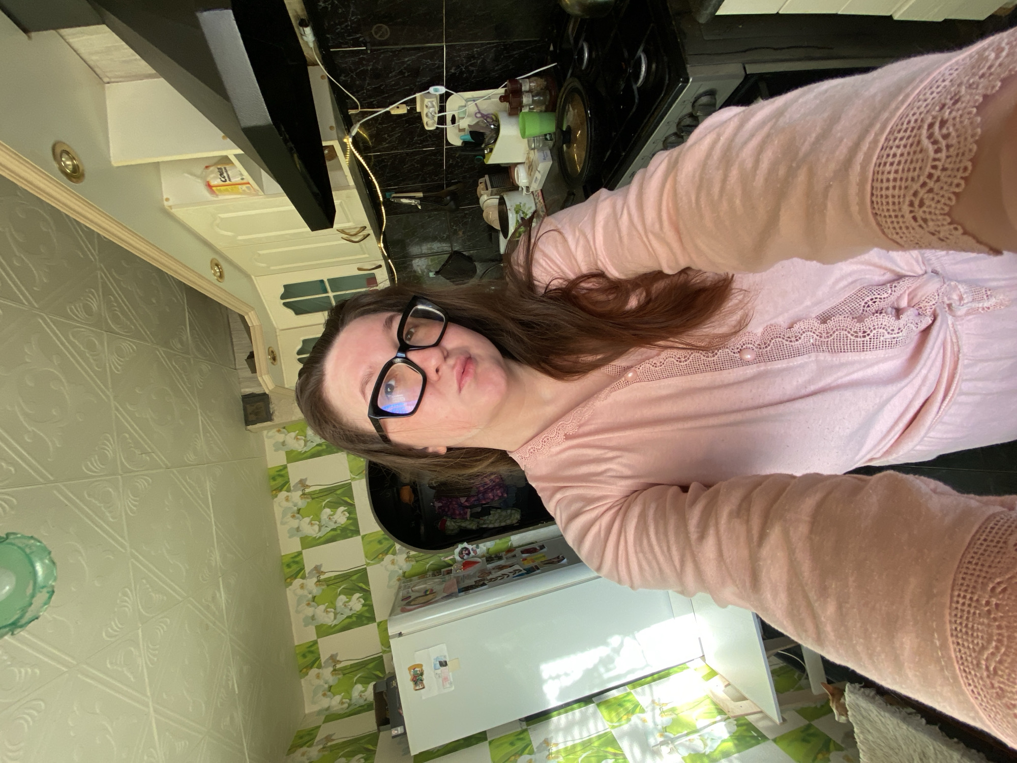 The image depicts a female individual taking a selfie in a kitchen located in Chaikovsky, Russia. The individual, with brown hair and black-framed glasses, is wearing a light pink long-sleeved top with lace detailing. She is looking directly at the camera with a slight puckering of the lips.
The kitchen features light-colored, embossed ceiling panels and walls with a patterned wallpaper depicting white orchids on a green and white checkerboard background. A section of the wall above the stove is covered in dark, possibly black, glossy tiles. Cream-colored upper cabinets with arched panels and gold-toned handles are visible, along with a black overhead range hood. A white refrigerator, adorned with magnets, stands on the left. A silver-colored gas stove is present, with a lidded pot resting on one of its burners. Various kitchen items, including a grater, mugs, utensils in a holder, and spice shakers, are arranged on the countertop. A packet labeled "Соль" (salt) is visible on an upper shelf. A string of small decorative lights is affixed to the wall below the upper cabinets. A window with horizontal blinds is partially visible on the far left, allowing sunlight to enter. An arched doorway or opening is visible to the right of the refrigerator, revealing hanging garments. A small orange cat is partially visible on the floor near a white furry rug, beneath a white table on the far left.