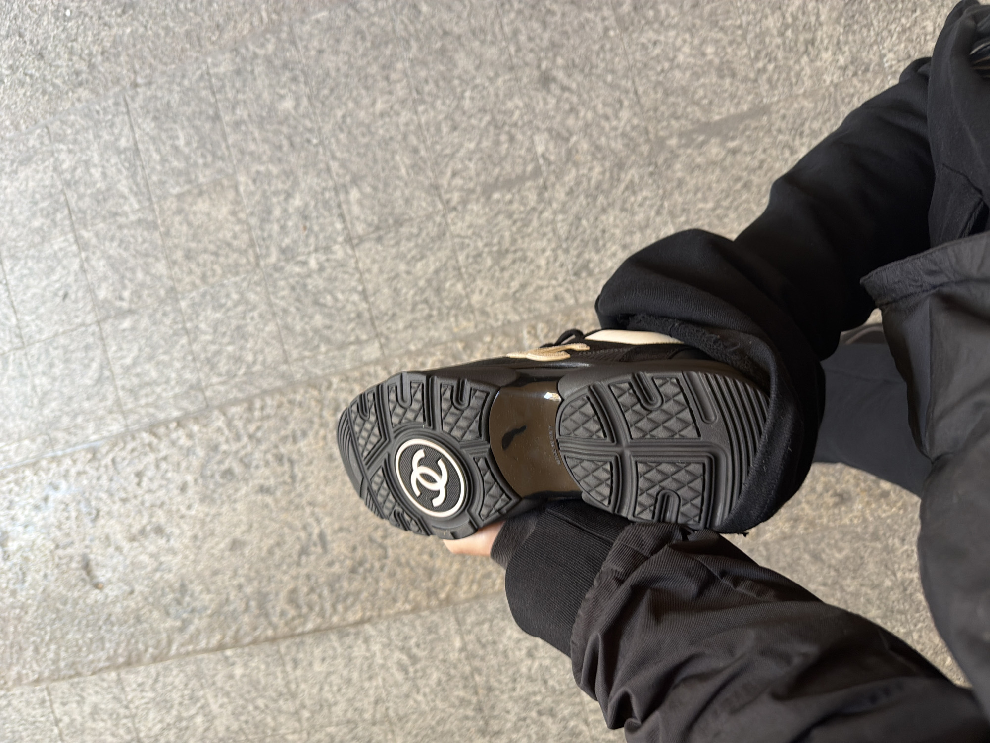 The image shows a person holding up a black Chanel sneaker, sole facing the camera, against a background of gray tiled pavement. The person is wearing a black jacket and sweatshirt. The sneaker's sole has the Chanel logo clearly visible. The location appears to be an urban area, likely Milan, Italy, due to the style of the pavement, though no specific landmarks are visible.
