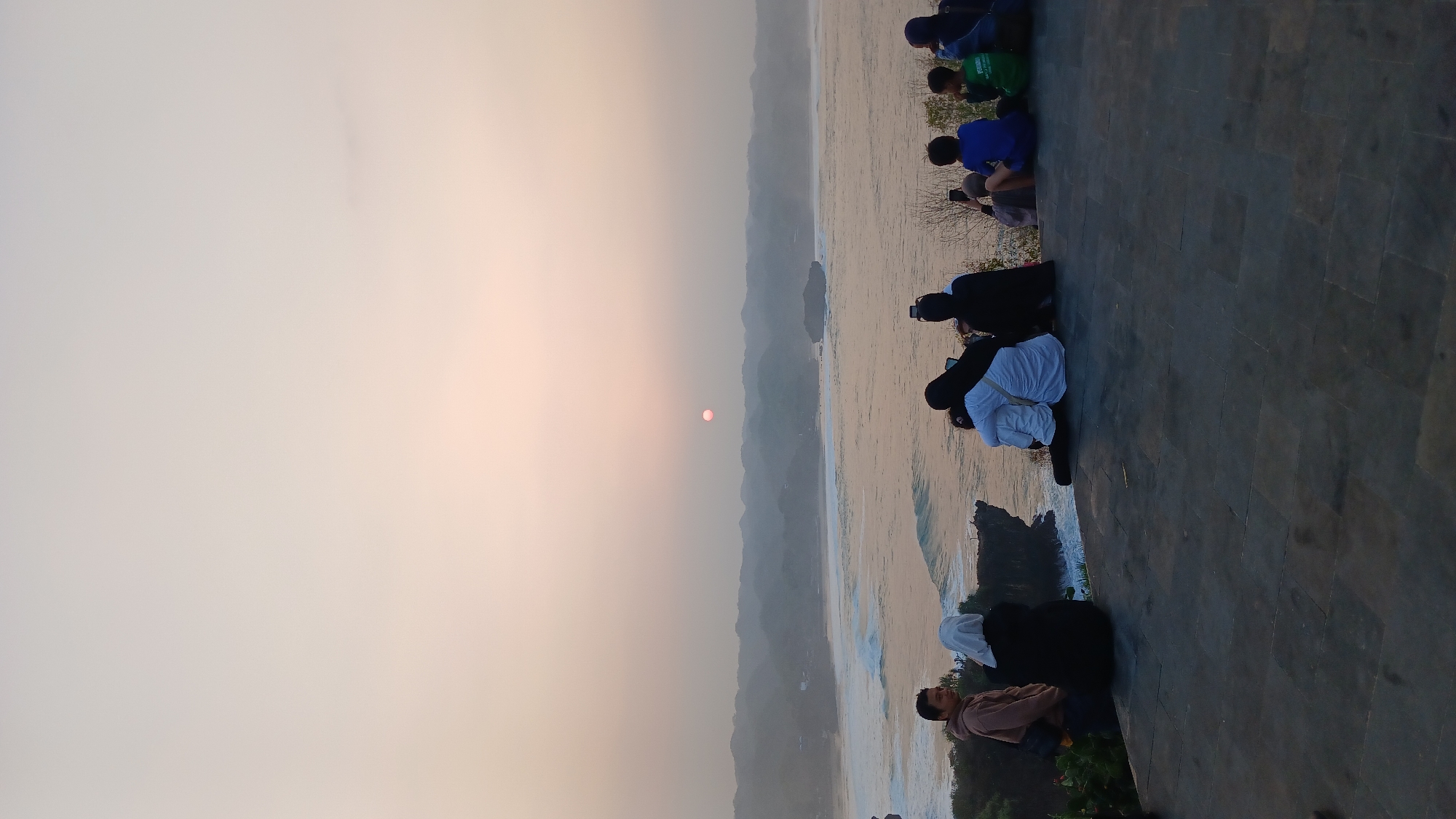 The image depicts a serene sunset scene at a coastal viewpoint in Tanjungsari, Indonesia. A group of approximately ten people are gathered on a paved cliffside overlooking the ocean. The sun, a pale pink orb, is low on the horizon, casting a soft, diffused light across the sky and the water. Gentle waves break on a sandy beach in the distance, and rolling hills form a hazy backdrop.
In the foreground, several individuals are seated and standing, their backs mostly turned to the camera. One man in a light brown hoodie stands to the left, looking towards the ocean. Next to him, a person in a black hijab and white shirt is seated. Further to the right, a couple is embracing while seated, with one person in a white shirt and the other in black. Another individual in a black hijab is seated next to them, looking at a phone. To the far right, a group of children and adults are seated, some facing the sunset and others looking down.
The ground is covered with dark, large square paving stones. The overall atmosphere is peaceful and contemplative, suggesting a popular spot for enjoying the natural beauty of the coastline at dusk.