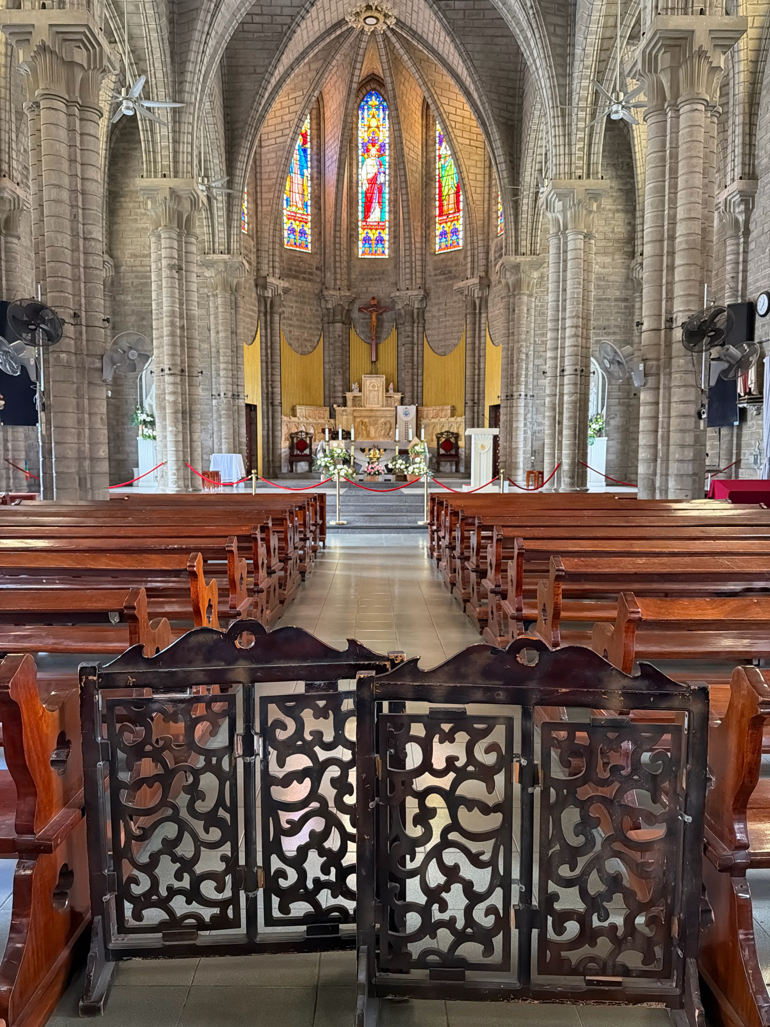 This image shows the interior of the Nha Trang Cathedral in Nha Trang, Vietnam. The cathedral is a large Gothic-style stone building with high vaulted ceilings and stained-glass windows. Wooden pews are arranged in rows on either side of a central aisle leading to the altar. In the foreground, two ornate wooden gates with intricate scrollwork are closed, blocking access to the pews. The walls of the cathedral are constructed of stone blocks, and there are several ceiling fans installed for ventilation. The altar area in the background features a cross, religious figures, and floral arrangements. The floor is tiled.
