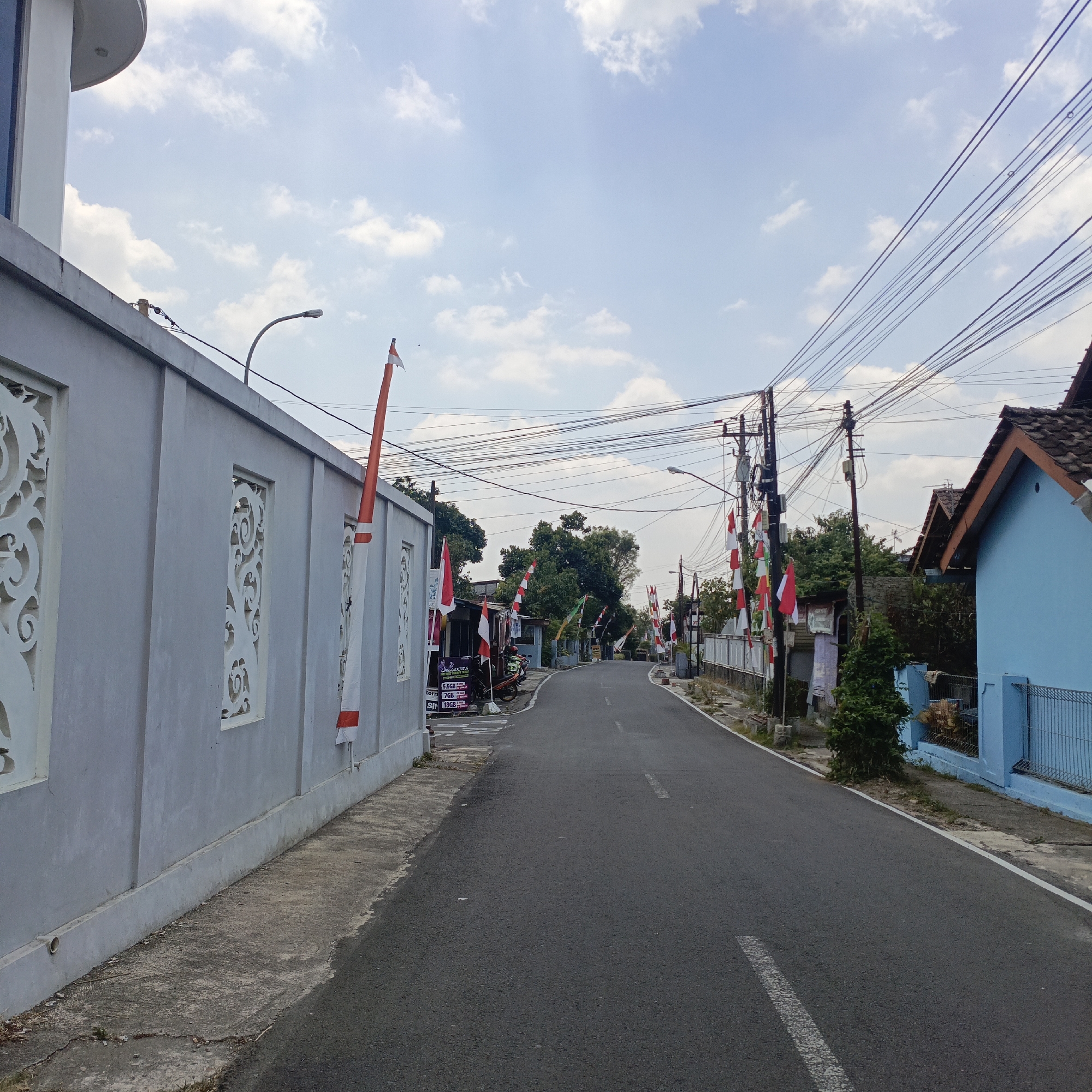 The image captures an outdoor scene of a paved road in a residential area. On the left side of the road is a light gray concrete wall with decorative cutouts. Several Indonesian flags, red and white, are displayed on poles along the wall and further down the road. Some of these flags are elongated pennants, while others are traditional rectangular flags.
The road itself is asphalt with white lane markings. On the right side of the road, there is a vibrant blue house with a tiled roof. A metal fence runs along the property line of the blue house. Power lines and utility poles are visible overhead, crisscrossing the sky. The sky is mostly blue with scattered white clouds.
In the background, there are trees and other houses, suggesting a suburban or semi-rural setting. There are no people clearly visible in the image, but the presence of flags implies a celebratory or patriotic occasion. The overall impression is a quiet street decorated for an event.