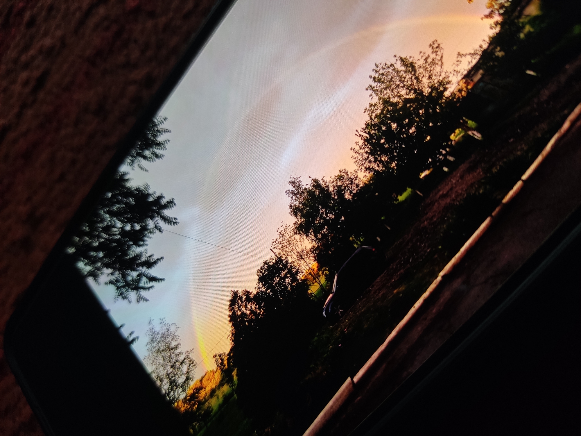 The image is a photograph of a cell phone screen showing a landscape scene. The landscape features trees, grass, and what appears to be a rainbow or atmospheric optical phenomenon in the sky. There's a vehicle, potentially a car, on the right side of the screen. The phone is photographed at an angle on a surface, which appears to be carpeted. The location is undetermined.