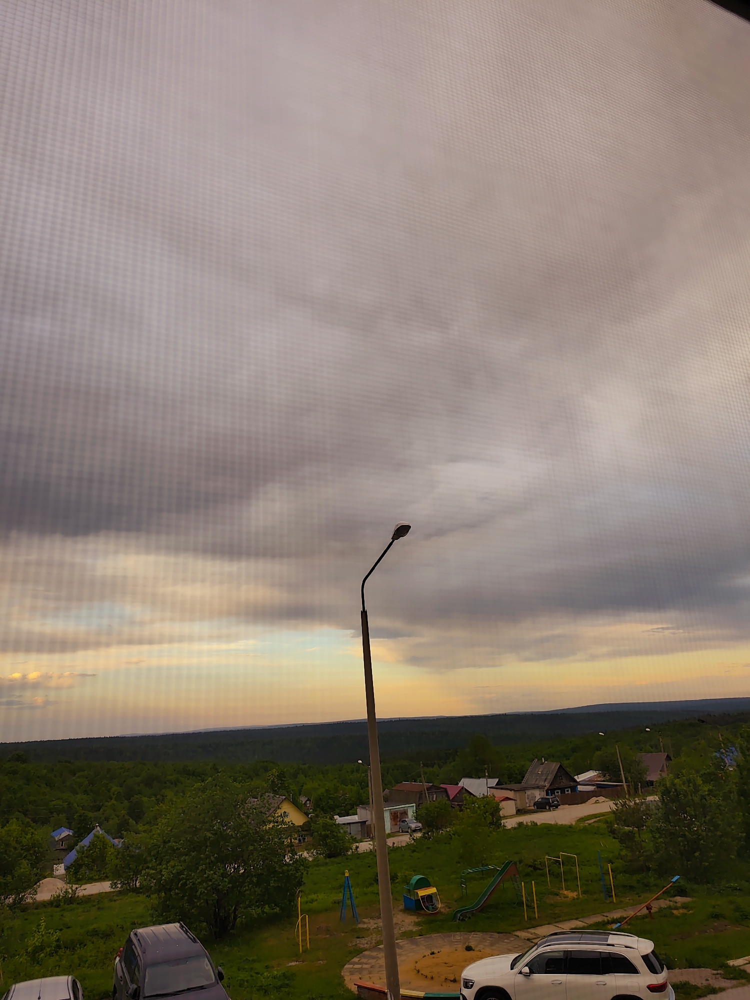 The image shows a view from a window in Gremyachinsk, Russia. The scene features an overcast sky with layers of grey and orange clouds. Below the horizon, a dense green forest spans the landscape. In the foreground, a residential area is visible, including several houses with varying roof colors (red, grey, and yellow). A playground with a slide and swings is located in the center of the frame. A black SUV and a white SUV are parked on the grass. A streetlight stands prominently in the center, partially obscuring the view.