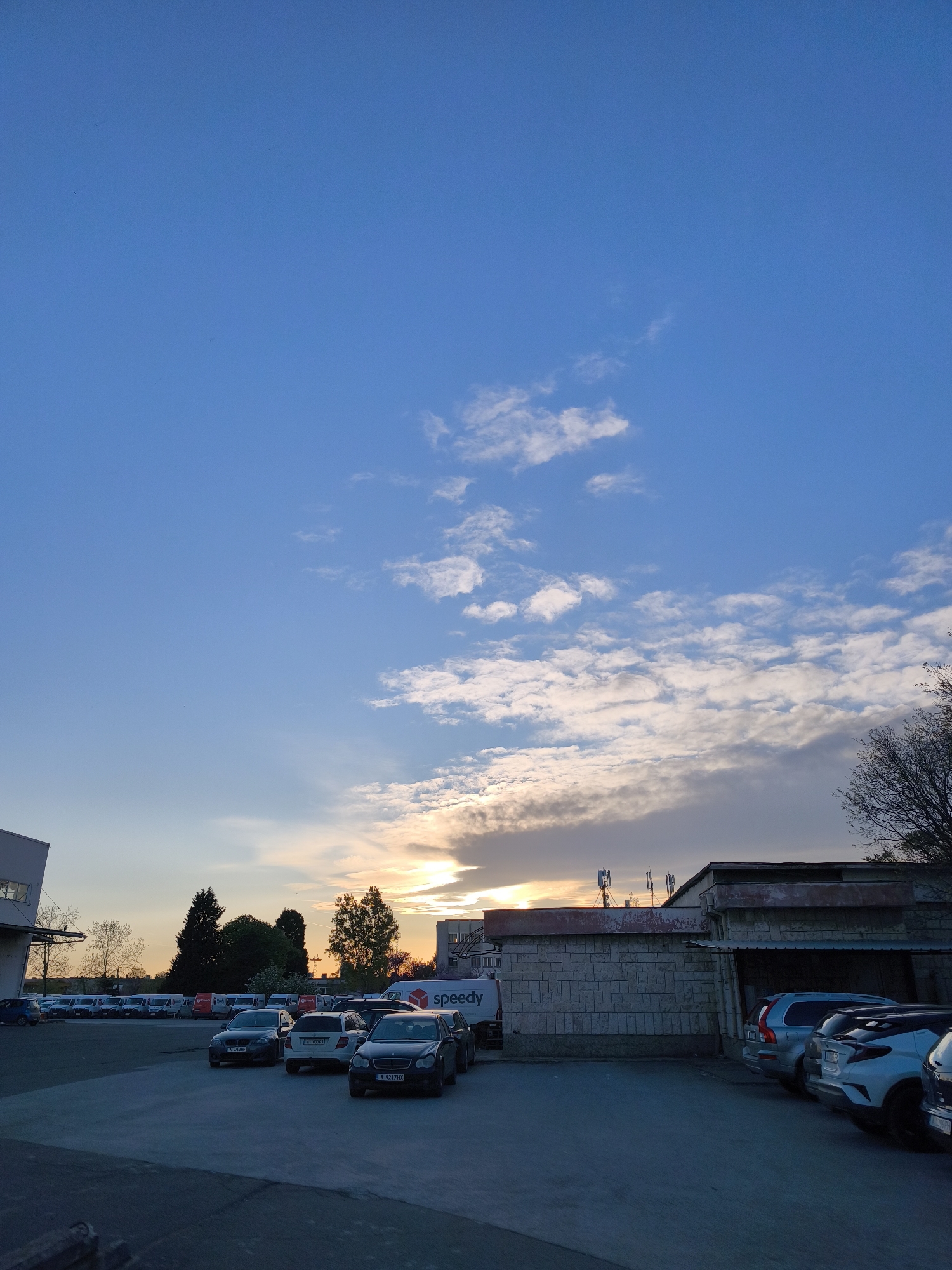 The image depicts a tranquil sunset scene over a parking lot and industrial area in Burgas, Bulgaria. The sky, a dominant feature, transitions from a clear, deep blue overhead to a lighter, more ethereal blue closer to the horizon, where the setting sun casts a warm, golden and orange glow through scattered clouds. White cumulus clouds are visible across the sky, with some heavier cloud formations near the horizon.
In the foreground and midground, a spacious concrete or asphalt parking lot is occupied by numerous parked vehicles. These include various cars such as sedans and SUVs, with visible Bulgarian license plates (e.g., A 7631P, A 1059PX, A 9217HX), which denote the Burgas region. To the left, a row of white delivery vans, several of which display the distinct "Speedy" courier service logo, are parked neatly.
Architectural elements include a low-slung, block-built structure on the right with a flat roof hosting several cellular antennas. A larger, more industrial-style building is partially visible on the far left. Trees with sparse branches, indicative of the season, are interspersed throughout the landscape, silhouetted against the colorful evening sky. The overall impression is one of calm and quiet at the close of day, with no people visible and no specific activity taking place beyond the vehicles being parked.