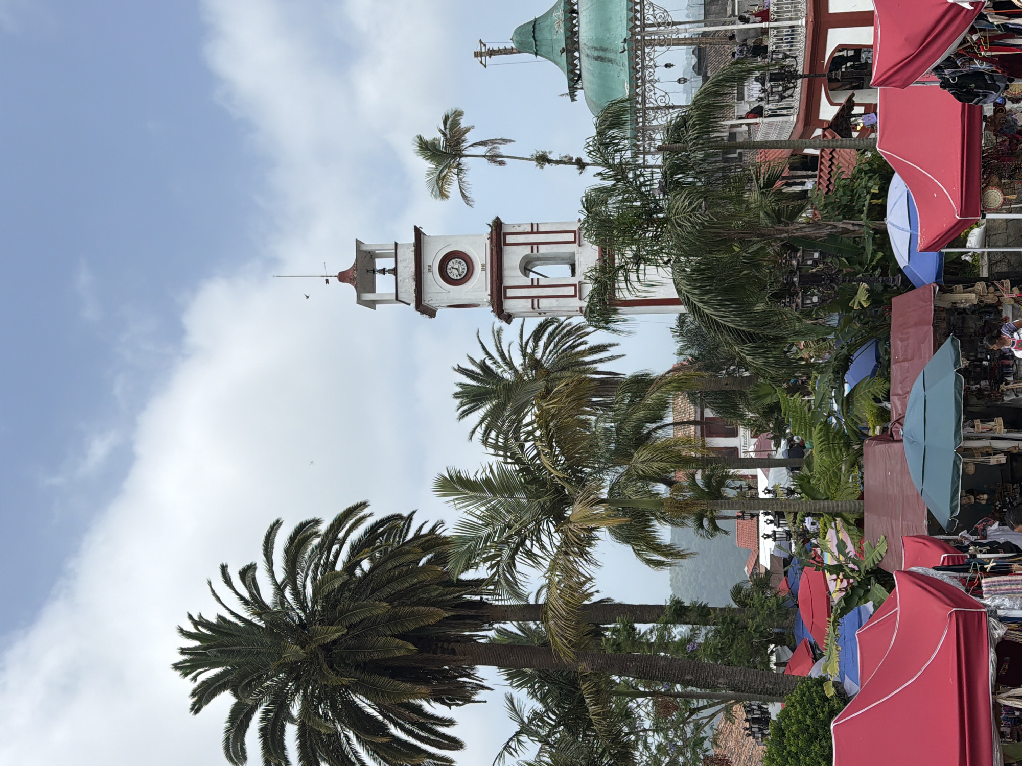 This image shows a clock tower and the green dome of the gazebo in Cuetzalan, Mexico. The buildings are white with red trim and situated among tall palm trees. Red and blue canopies of market stalls appear in the foreground on the right. The sky is blue with some clouds.