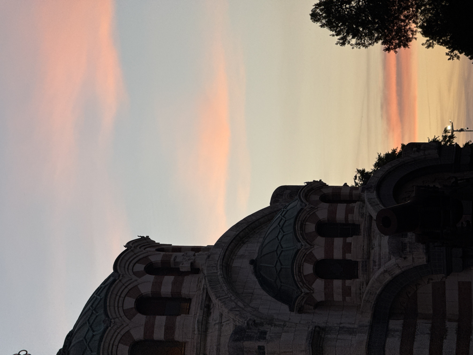 A low-angle, vertically-oriented shot shows the Alexander Nevsky chapel mausoleum in Pleven, Bulgaria, at twilight. The structure's domed roofline, ornamented with red and white banding and multiple arch-shaped windows, occupies the lower right quadrant. The silhouette of foliage occupies the top right corner of the image. The sky above the structure transitions from a pale grey in the lower left to peach and yellow hues near the horizon. Narrow, pink-toned clouds stretch vertically in the upper portion of the shot. The light pole and trees in the background cast a silhouette near the right top side.