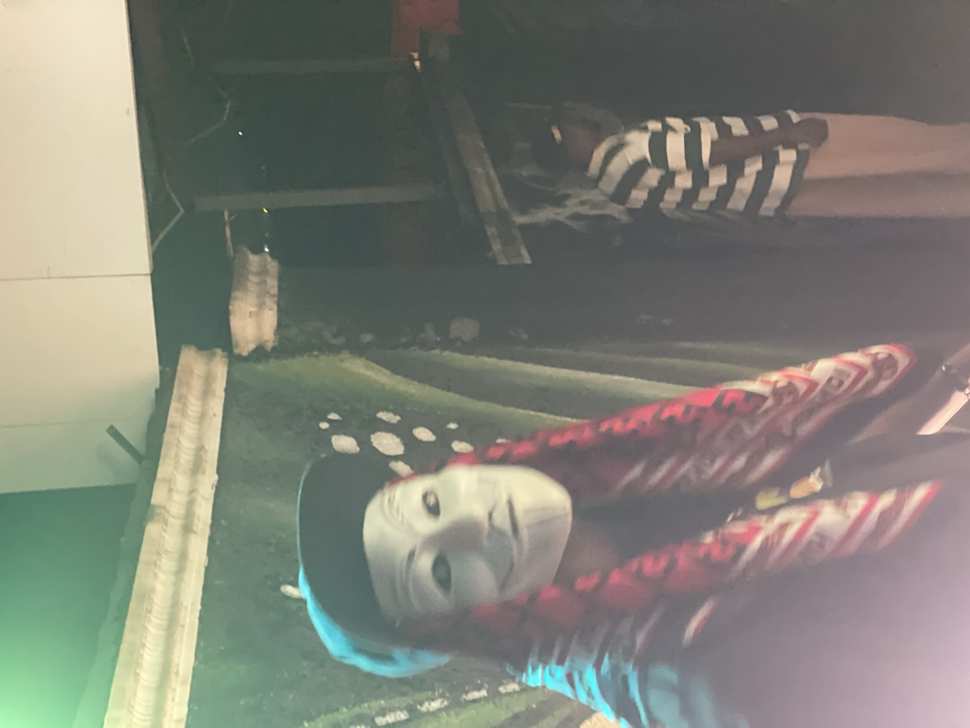 In Kurunduma, Nigeria, a person wearing a white Guy Fawkes mask and a black baseball cap is in the foreground. Their hair appears to be red braids, and they are wearing a patterned scarf around their neck. Behind them, another person is visible, wearing a striped shirt and light-colored pants. The background suggests an outdoor setting with some structures, possibly under construction or in disrepair, with a dark, moody ambiance.