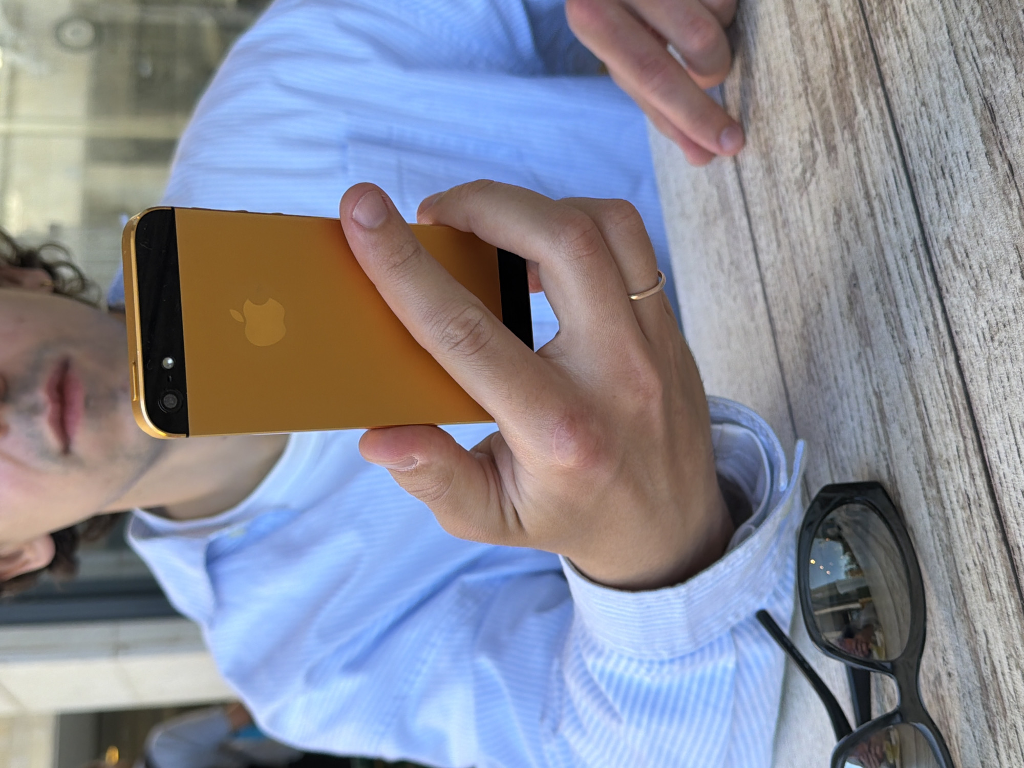 The image presents a close-up, rotated view of a person holding a gold-colored smartphone. The subject, a male with dark, curly hair, is partially visible on the left, wearing a light blue button-up shirt with subtle white stripes. His right hand holds the phone, while his left hand rests on the table.
The smartphone, an iPhone model, is distinctively gold, featuring a black segment at its top back that houses the camera lens and flash. The iconic Apple logo is clearly visible and also gold-colored on the phone's back panel. A slender gold-colored ring adorns the ring finger of the hand grasping the phone.
On the light, weathered wooden table to the right of the person's hand, a pair of black sunglasses with reflective lenses is placed. The background suggests a bright, possibly outdoor setting, such as a cafe or restaurant, fitting the provided context of Tel-Aviv, Israel. The person's face is largely out of focus, and no specific emotions are discernible. The only visible text is the Apple logo on the phone.