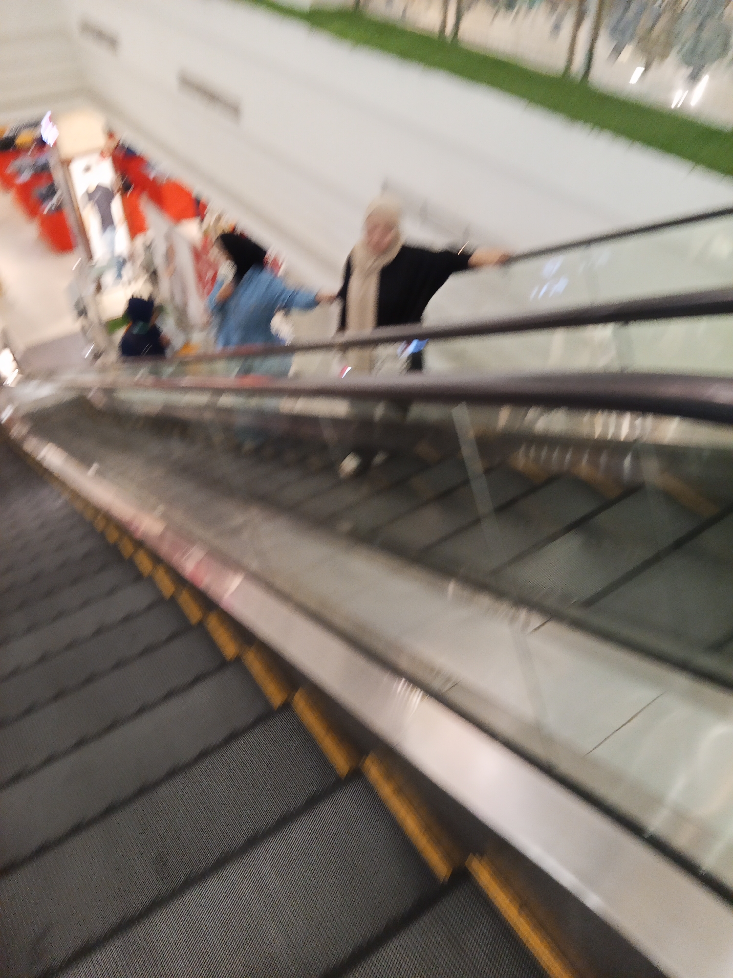 The image shows an interior scene of a shopping mall or department store in Lubuklinggau, Indonesia. The primary focus is on an escalator, captured in motion, with several individuals on it. On the upper landing of the escalator, a person wearing a light-colored hijab and a dark top is visible, leaning on the handrail. Below this person, on the escalator steps, another individual in a bright blue garment is also using the escalator. Further down the escalator, another person in dark clothing is seen. The escalator itself is dark gray with yellow safety markings. The surrounding environment appears to be a brightly lit interior with white walls and some hints of displays or merchandise on the upper level. The overall image is slightly blurred, indicating movement.