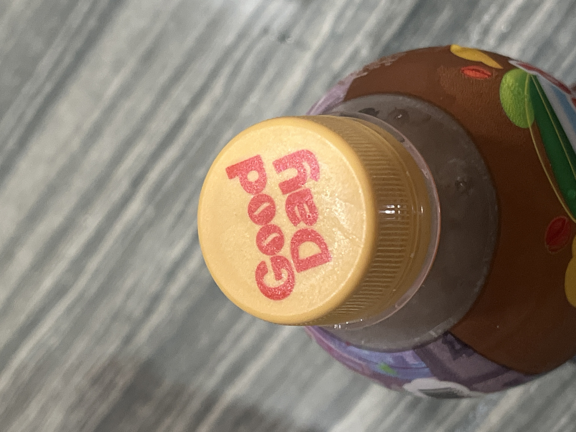 A close-up, overhead shot focuses on the cap of a brown beverage bottle. The cap is yellow and features the red text "Good Day" in a playful font. The bottle itself is brown and appears to have colorful graphics around its base, though these are only partially visible. The background consists of a textured, gray surface with diagonal lines, possibly a wooden table. The scene is devoid of people, actions, or interactions. The location in Teluk Air, Indonesia, is not discernible from the visual information provided.