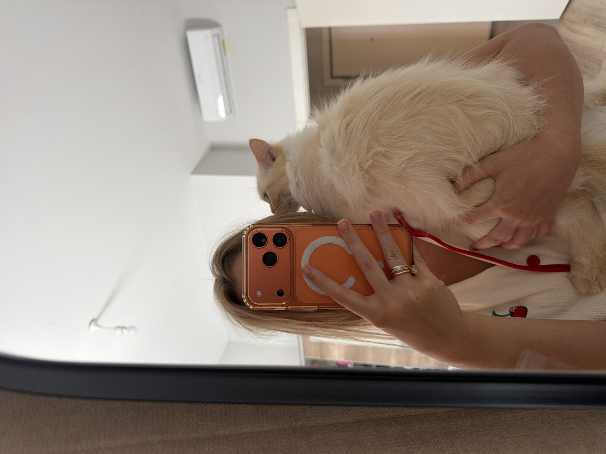 A person, whose face is obscured by an orange-cased smartphone they are holding, is taking a mirror selfie while holding a light-colored cat. The person is wearing a white top with a red cherry embroidery and red trim, and a red lanyard. The cat is light cream-colored with darker points on its ears and face, and is nestled against the person. The background shows a bedroom with a white wall, a ceiling-mounted air conditioning unit, a light fixture, and a doorway. A large mirror with a dark frame is on the left side of the image, reflecting the scene. The overall impression is of a personal moment captured in a domestic setting.