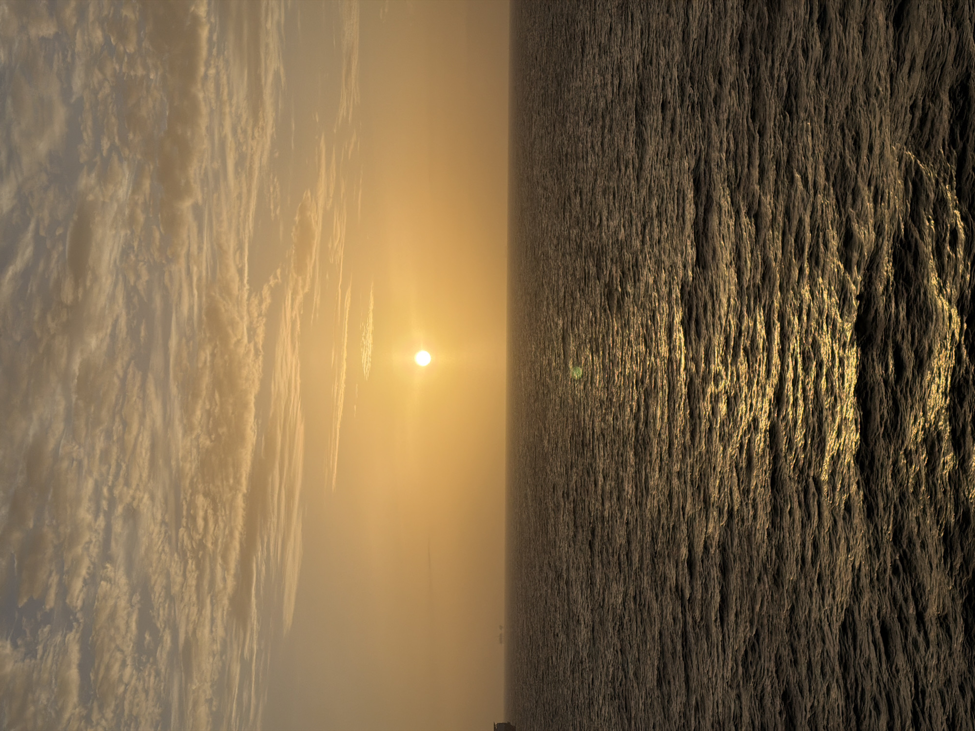 The image depicts an ambiguous scene split vertically. The left side shows a sky filled with striated clouds, illuminated with a golden hue from the setting sun. The sun, a distinct bright disc, is positioned centrally, creating a halo effect. The right side shows a water surface with intricate wave patterns, reflecting sunlight. The water texture appears rough and textured. The horizon line is not clearly defined, suggesting either a close-up perspective or a diffused atmospheric condition. The location is not discernible from the image, and there are no visible signs of human activity.
