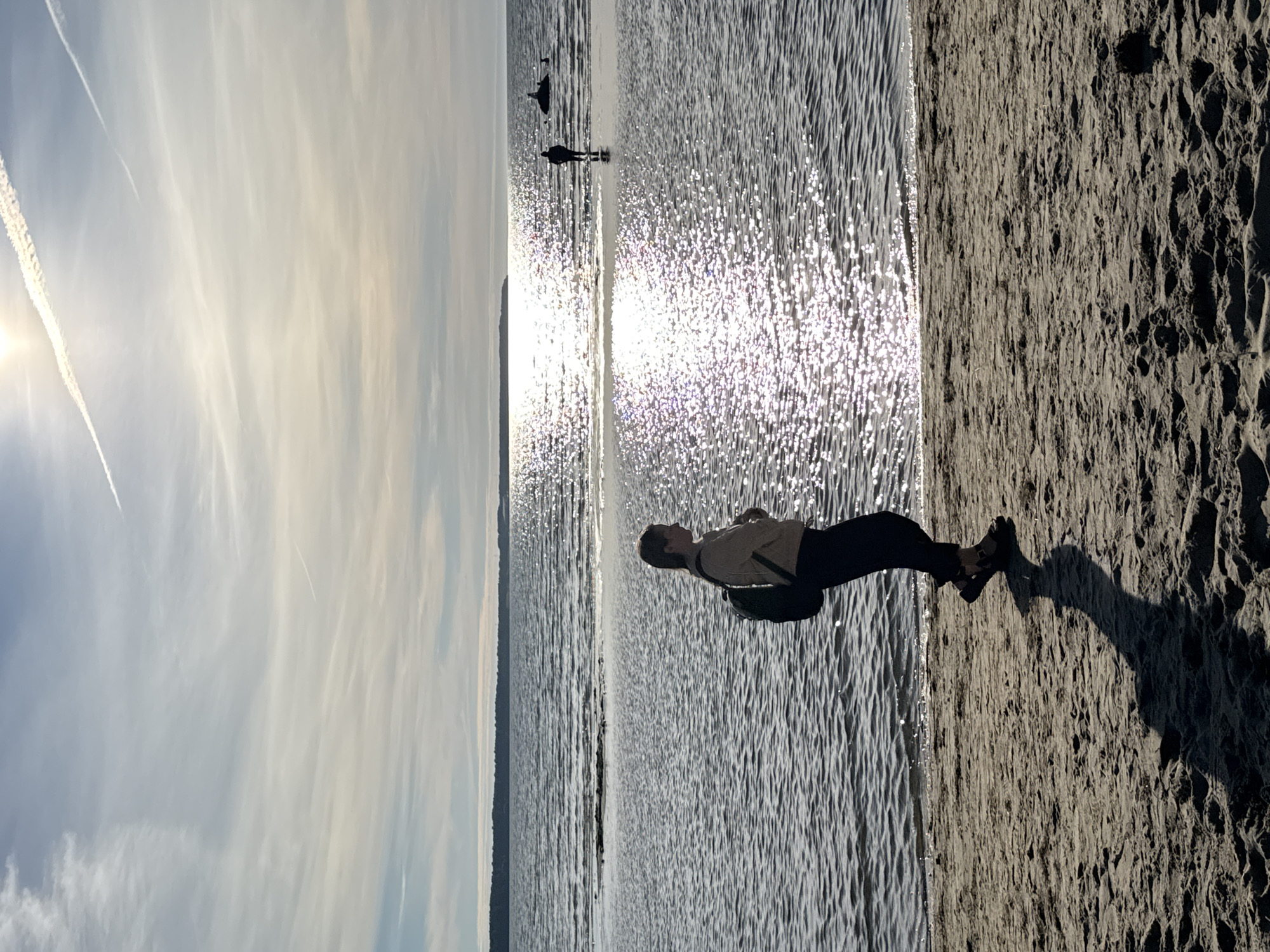 A silhouetted person walks along a sandy beach in Tallinn, Estonia, with the Baltic Sea stretching out before them. The sun is low in the sky, casting a bright, shimmering reflection on the water. Another person and a small boat are visible in the distance, wading in the shallow water. The sky is a mix of blue and white clouds, with contrails from an airplane visible. The sand is textured with footprints and debris. The overall mood is one of a peaceful, late afternoon by the sea.