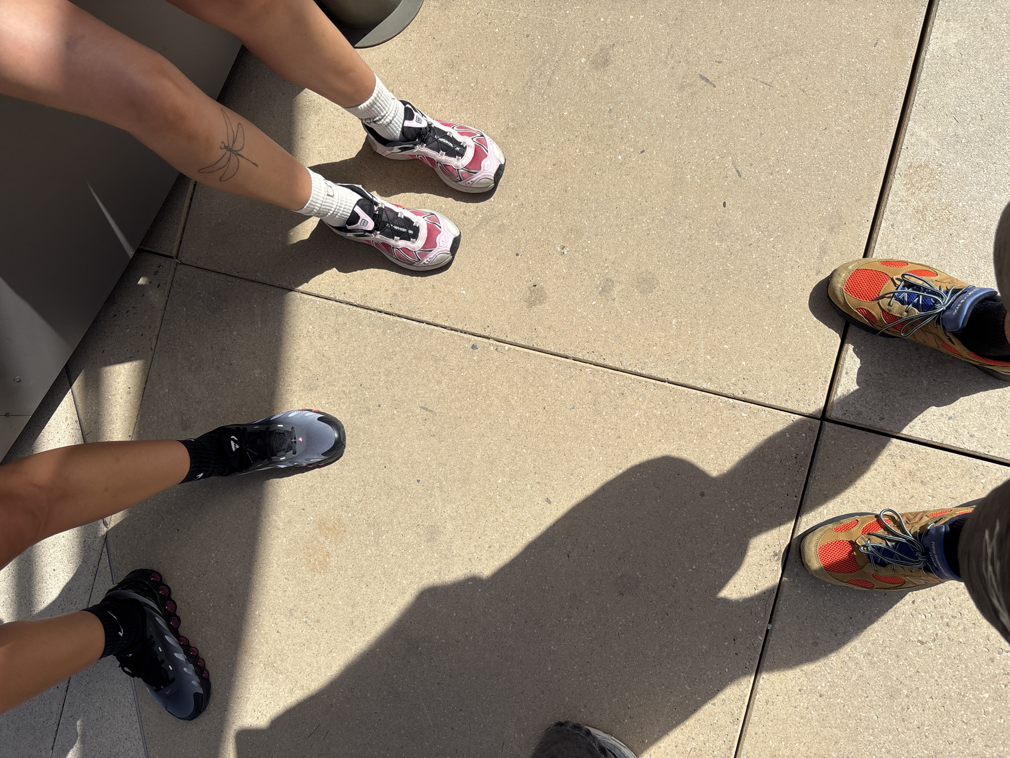 The image shows the lower legs and feet of at least three individuals standing on a light-colored tiled surface, possibly a public walkway in Barcelona, Spain. Sunlight casts strong shadows on the ground. One person wears dark grey and maroon sneakers with black socks. Another person, whose right leg is partially visible, has a dragonfly tattoo on their calf and wears pink, white, and black athletic shoes with white socks. A third person is wearing tan, red, blue, and black athletic shoes, also with dark socks. The tiled surface appears to be made of large square tiles. In the background, a dark, angled surface suggests a building or structure. The overall scene suggests an outdoor urban environment.