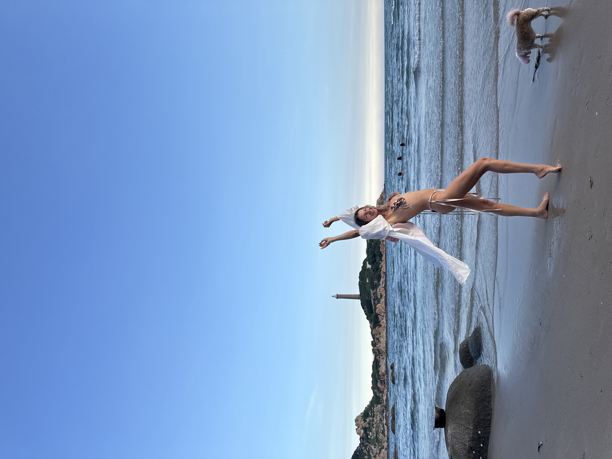 In the city of Thuận Thành, Vietnam, a woman in a bikini stands on a sandy beach near the ocean. The woman is posing with her arms raised and a sheer white garment draped over her. A small, light-colored dog is on the beach near her. In the background, the ocean stretches to the horizon, with a lighthouse visible on a rocky outcrop in the distance. The sky is clear and blue.