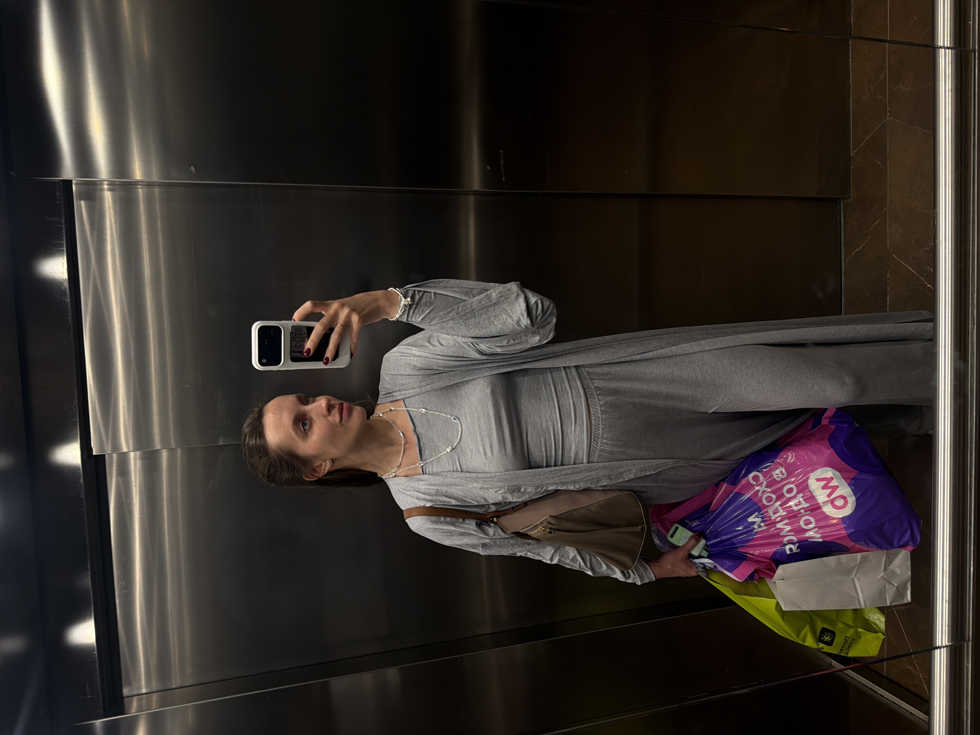 A woman is taking a selfie in an elevator. She is wearing a gray outfit and has a shoulder bag and shopping bags in her hands. One of the shopping bags has Cyrillic writing on it and the letters "GW". The elevator has stainless steel walls and a tiled wall. The text on the shopping bag reads "МОЛОДЦЫ, ДОБРО ПОЖАЛОВАТЬ", which is a Russian phrase that translates to "Well done, welcome." The GW logo is for "Gulliver World," which is a chain of toy stores in Russia, located in Moscow.