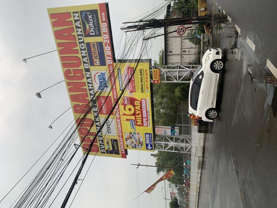The image captures a street scene in Singosari, Indonesia, at the coordinates -7.914 latitude, 112.656 longitude. A white car is parked on the right side of the street, facing towards the camera. The street is wet, suggesting recent rain.  A large billboard on the left side of the street advertises "SUPERMARKET BARAHAM BANGUNAN" with several brands like "JOTUN", "DULUX", and "TOTO". A smaller billboard beneath the main one, is advertising "GERAI MALANG" and "DEPO BANGUNAN". The scene also includes multiple electrical wires running across the street. There are trees and vegetation on the left side of the street. The scene appears to be captured on a cloudy day, with a gray sky in the background.  A red flag is visible behind the parked car.  A sign with the word "MASUK"  (which translates to "Enter") can be seen on the billboard.  There is a man, potentially a worker, in an orange shirt visible behind the car, possibly working on the billboard.