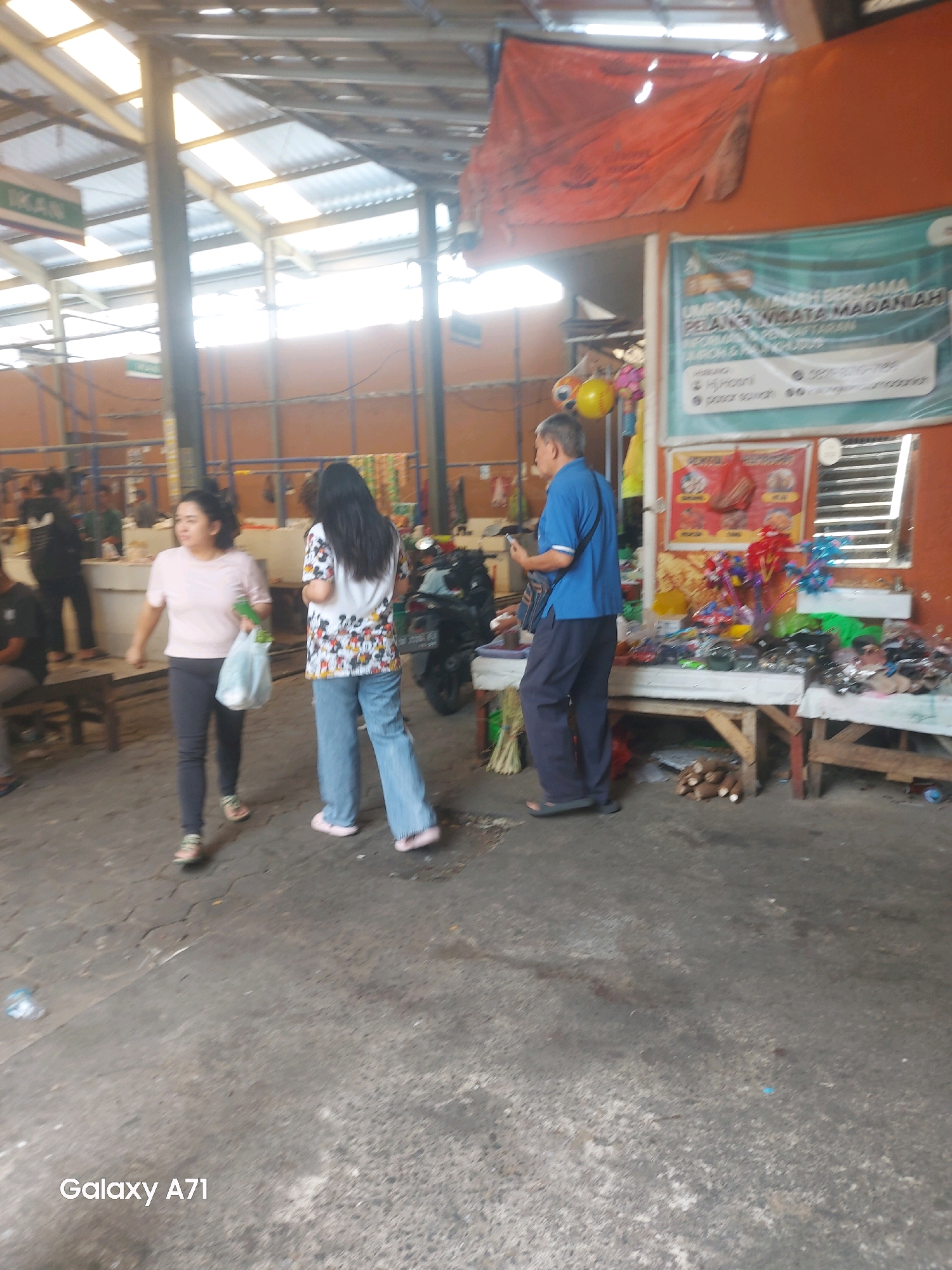 The image depicts a bustling market scene, likely in Makassar, Indonesia, given the presence of "Galaxy A71" watermark, a Samsung phone model popular in the region. In the foreground, a woman in a pink shirt and dark pants walks carrying a plastic bag. To her right, another woman with long dark hair wears a colorful Mickey Mouse t-shirt and light blue jeans. Behind them, a man in a blue polo shirt and dark pants stands looking at his phone. The market is covered by a corrugated metal roof supported by metal beams. Stalls display various goods, including toys and packaged items. A motorcycle is parked behind the women. The ground is paved with concrete, showing signs of wear and dirt. The overall atmosphere suggests a lively, everyday commercial environment.
