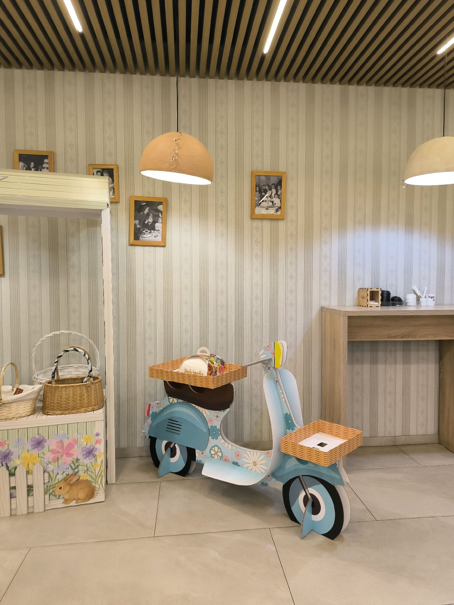 The image depicts an indoor scene that appears to be a retail or decorative display, possibly in Chișinău, Moldova. The focal point is a life-sized, turquoise cardboard cutout of a vintage scooter, adorned with floral patterns. The scooter has two wicker baskets, one on the front handlebar and another on the rear rack, both appearing to hold small items. To the left of the scooter is a white wooden display stand holding several wicker baskets, one of which has a fabric scarf. In front of the display stand is a white wooden box decorated with painted flowers and a rabbit.
The background features a wall with a subtle striped wallpaper pattern. Several framed black and white photographs are displayed on the wall, depicting groups of people in what appear to be formal settings. Two large, dome-shaped pendant lights hang from the ceiling, which has a slatted wooden design with integrated lighting. To the right, a light-colored wooden counter or desk is visible, with some small wooden boxes and stationery items on its surface. The flooring is a light-colored tile. No people are present in the scene.
