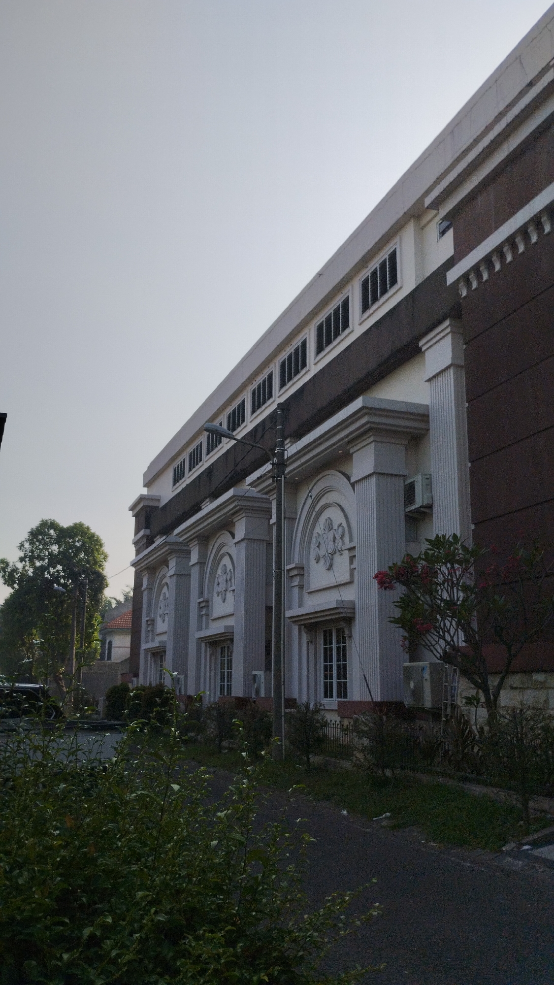 The image displays the exterior of a modern building with classical architectural elements. The building has a light-colored facade featuring prominent columns and arched decorative motifs above windows. The upper section of the building is darker, with a band of evenly spaced dark windows. A streetlamp is visible in front of the building. The surroundings include greenery, with trees and bushes in the foreground and to the left. The sky is clear and light. A paved path or road runs alongside the building. The architecture suggests it could be a public building, institution, or office complex. The presence of air conditioning units on the facade indicates a warm climate. The image captures the building from a low angle, emphasizing its scale.