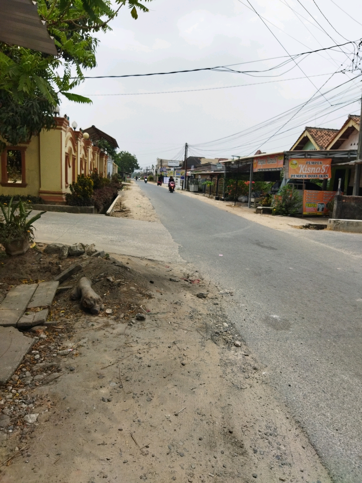 A street scene in Banjar Agung, Indonesia, shows a two-lane asphalt road with a sandy and rocky shoulder. On the left side of the road, a building with a yellow facade and arched windows is visible, bordered by a low wall. Adjacent to this building is a small trench. Further down the street on the right, several small shops are set up, including one with an orange banner displaying "PEMPEK Kisna87" and "PEMPEK DOSS IKAN." A few individuals on motorcycles are traveling down the road, and overhead, a dense network of electrical wires crisscrosses the sky. The environment appears to be moderately populated with a mix of residential and commercial structures.