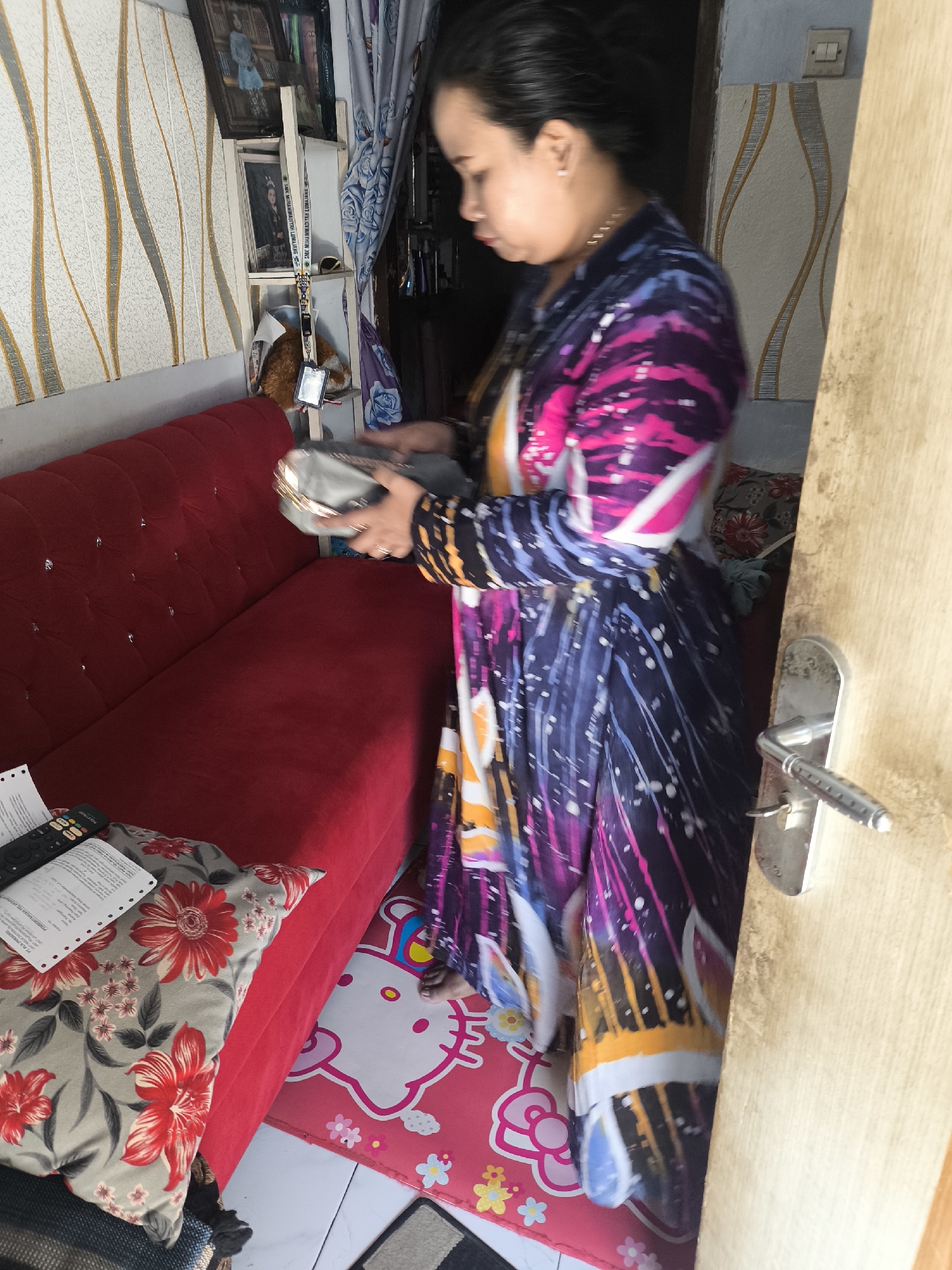In a room in Labruk Kidul, Indonesia, a woman wearing a colorful, patterned dress is standing near a doorway. She is holding a silver clutch bag in her hands, which are positioned in front of her. The room features a red sofa with tufted details and a decorative pillow with red floral patterns. A floor mat with a Hello Kitty design is visible on the floor. In the background, there is a shelf with framed pictures and a curtain with a floral pattern. A door handle is visible on the right side of the image.