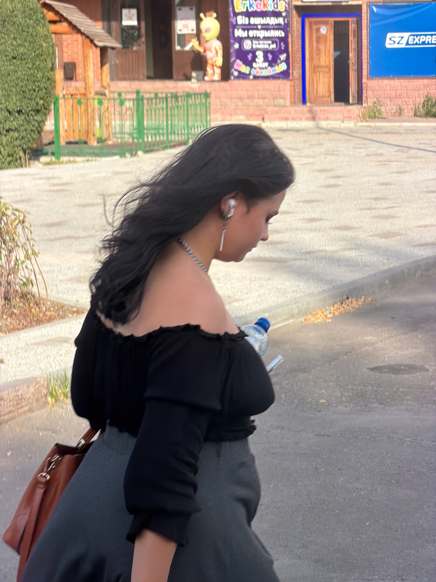A young woman with long, dark, wavy hair, wearing a black off-the-shoulder top and a gray skirt, walks away from the camera. She has on white earbuds and a silver necklace with a pendant. She is holding a clear plastic water bottle with a blue cap in her right hand. A brown leather shoulder bag is slung across her left shoulder. She is walking on an asphalt surface, with a concrete curb and a patch of dry leaves and debris to her right. In the background, there is a building with a brick facade, a wooden door, and a blue banner with white lettering that reads "SZ EXPRESS". To the left of the building, there is a green metal fence and a small wooden structure with a brown roof. There is also a tall, dense green hedge on the far left of the image. The overall scene suggests an outdoor urban environment. The presence of the "SZ EXPRESS" banner and the architectural style of the building, combined with common visual elements in Pavlodar, Kazakhstan, suggests this location.