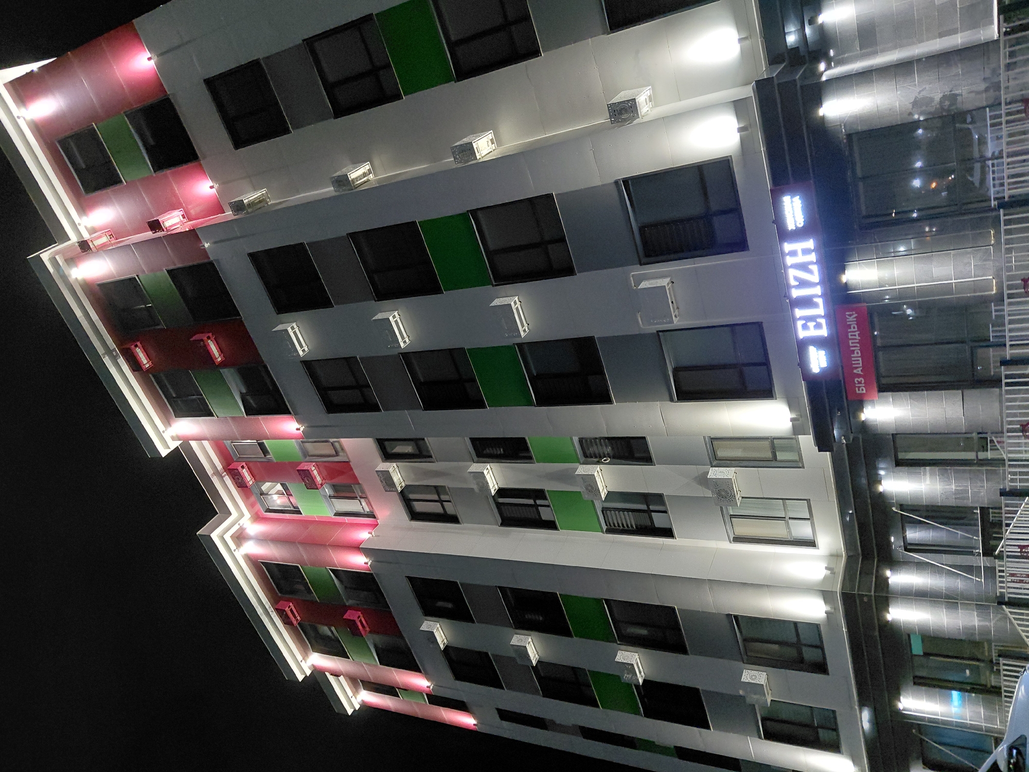 The image shows a modern, multi-story building at night in Aktau, Kazakhstan. The building has a distinctive design with alternating bands of white, green, and red. It is illuminated by a combination of warm white and pink lights, highlighting its architectural features and creating a vibrant aesthetic. Multiple windows are visible on each floor, some with blinds. Several outdoor air conditioning units are mounted on the exterior walls. At street level, a business with a prominent neon sign reading "ELIZH" is visible, along with text in Kazakh script below it. The building is fronted by a metal fence and stairs leading to the entrance. The sky above is dark, indicating nighttime.