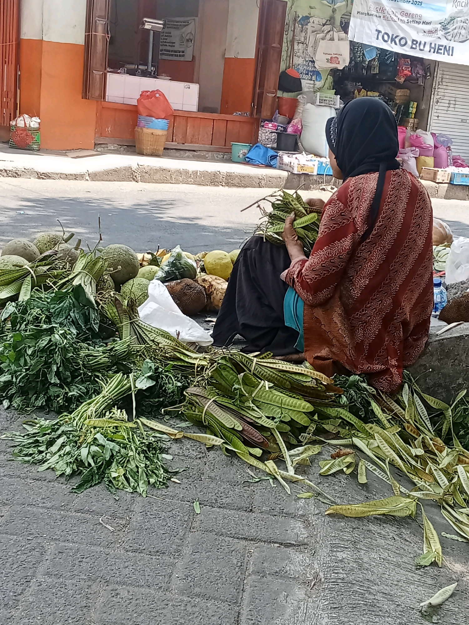 In Sukowinangun, Indonesia, a person wearing a dark headscarf and a patterned red and white robe is sitting on a concrete ledge, facing away from the camera. They are holding a bunch of green, leafy vegetables in their hands, with more vegetables and some larger, round fruits piled around them on the ground. The scene appears to be an outdoor market, with a building and shopfront visible in the background. The ground is paved with textured gray concrete.