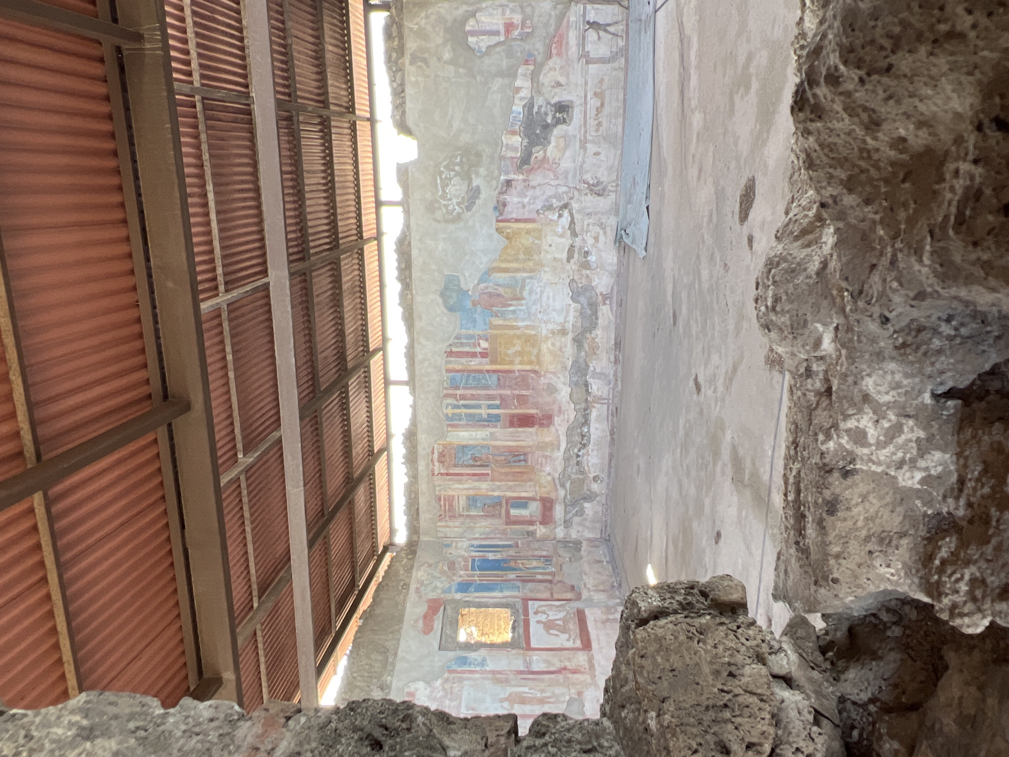 This is an image of a partially preserved room in Pompeii, Italy, featuring ancient Roman frescoes. The room is covered by a modern corrugated metal roof supported by wooden beams, suggesting preservation efforts. The walls display vibrant, though faded, frescoes with figures and architectural motifs in red, yellow, blue, and white. Some areas of the frescoes are damaged or missing. The floor is bare earth or concrete. Large stones and rubble are visible in the foreground, framing the view of the room. The light suggests it is daytime.