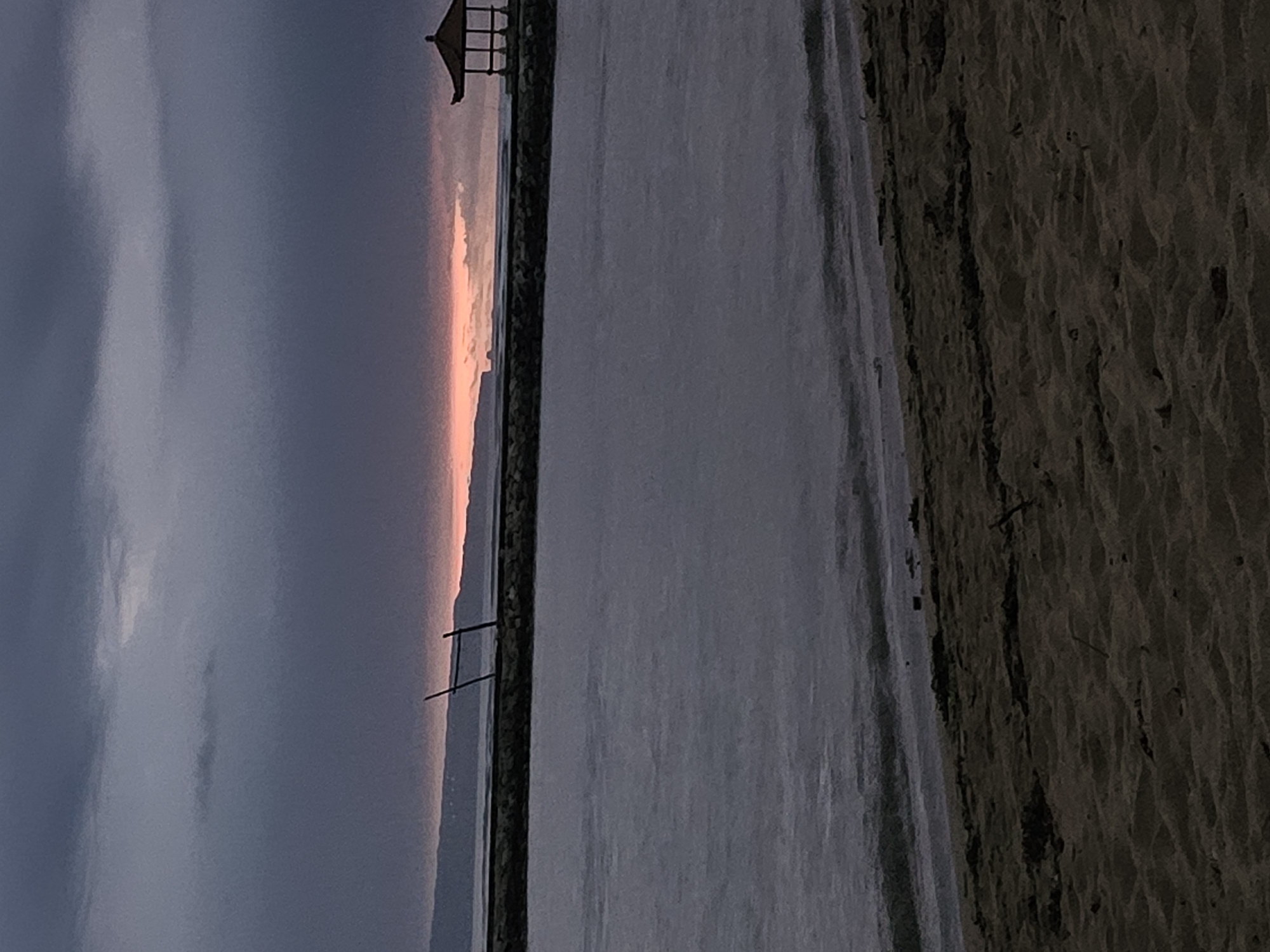 The image depicts a dusk scene at a beach in Denpasar, Indonesia. The sky is cloudy with a band of orange light on the horizon. A breakwater separates the sky from the sea, reflecting the sky's colors. The beach is visible on the right side of the image. A small gazebo sits atop the breakwater in the distance.