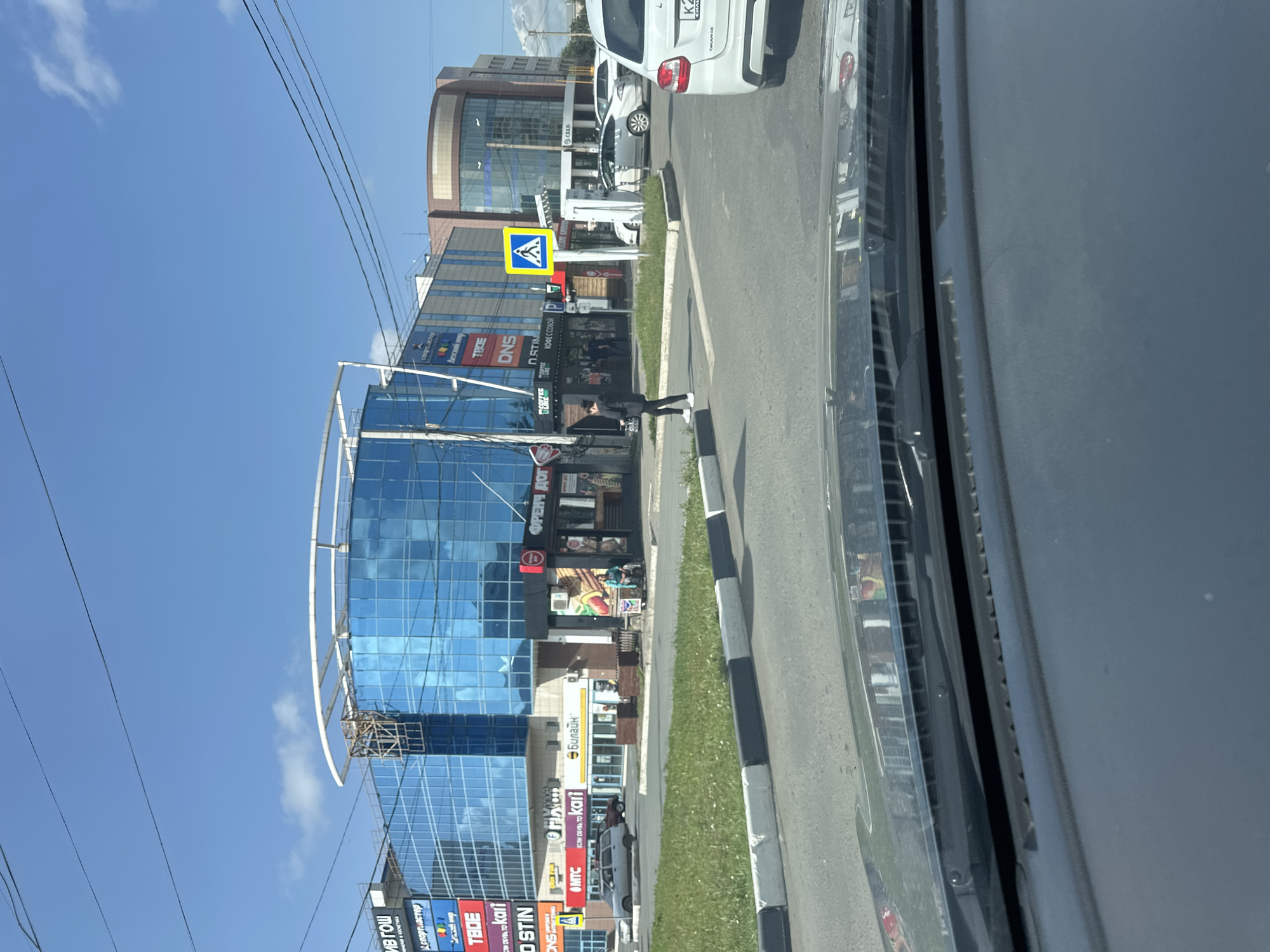 The image captures a street scene in Orsk, Russia, with a large, modern shopping center as the focal point. The shopping center features a distinctive blue glass facade and a white curved roofline. Signs on the building indicate various businesses, including "Спортмастер" (Sportmaster), "Детский мир" (Children's World), and "DNS". A standalone kiosk with the sign "Френч дог" (French Dog) is also visible.
A pedestrian, dressed in a black suit and white sneakers, is crossing a street in front of the shopping center, utilizing a marked crosswalk. A pedestrian crossing signal, a blue sign with a white silhouette of a person walking, is positioned on the right side of the crosswalk. Several cars are on the road, including a white sedan with a license plate starting with "K2" on the right side of the image. The environment appears to be a sunny day with a clear blue sky and some scattered clouds. The foreground shows the interior of a vehicle, suggesting the photo was taken from a car.