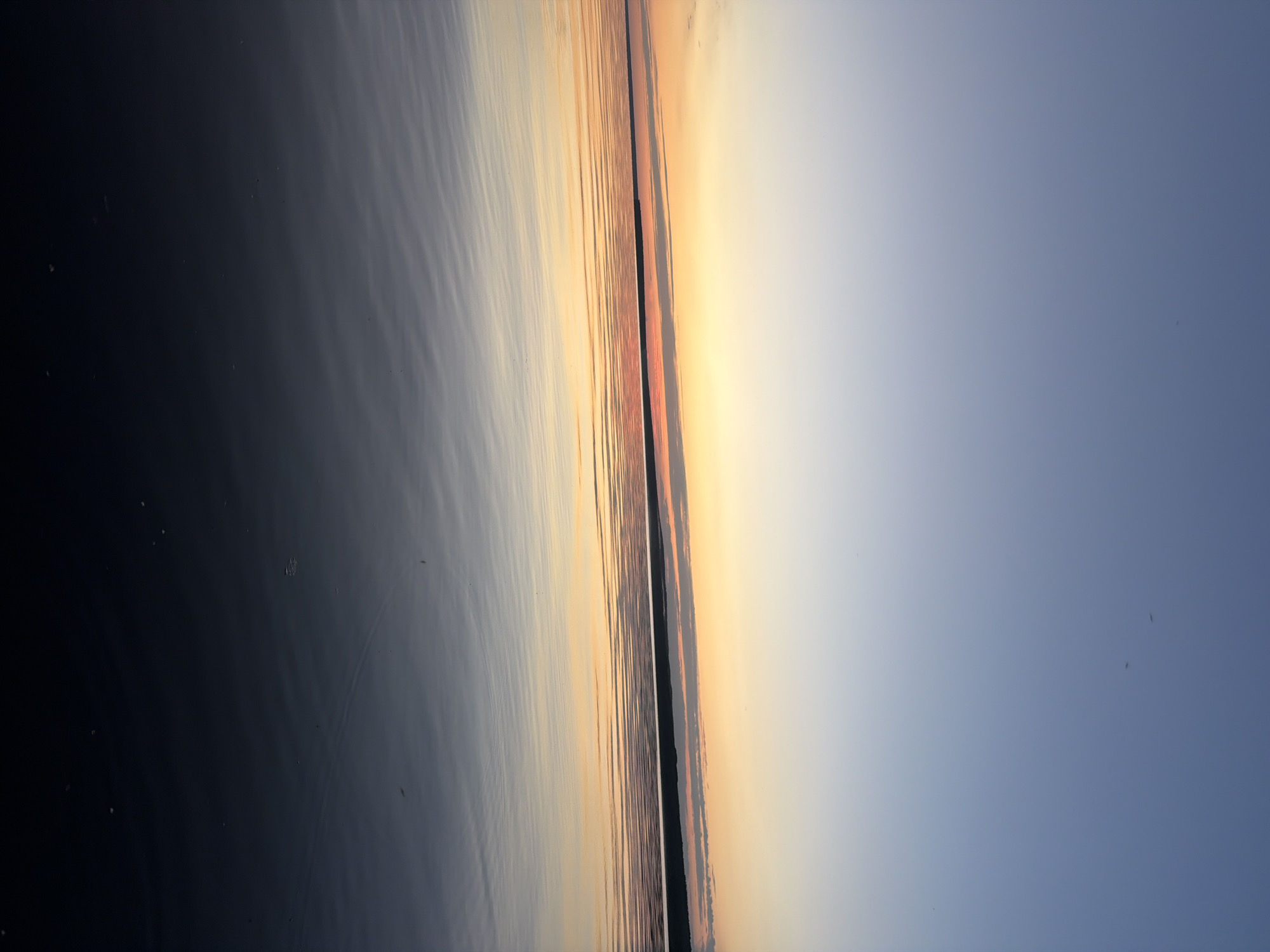 The image depicts a serene sunset over the Kama River in Chaikovsky, Russia. The sky transitions from a pale blue at the top to a warm gradient of yellow and orange near the horizon, where a dark, silhouetted treeline marks the distant shore. The calm surface of the river reflects the colors of the sky, creating a mirror-like effect with gentle ripples disturbing the smooth surface. The foreground is dominated by the dark, deep water of the river, with subtle reflections of the sky visible. No people or man-made objects are prominently visible, emphasizing the natural beauty of the landscape at dusk.