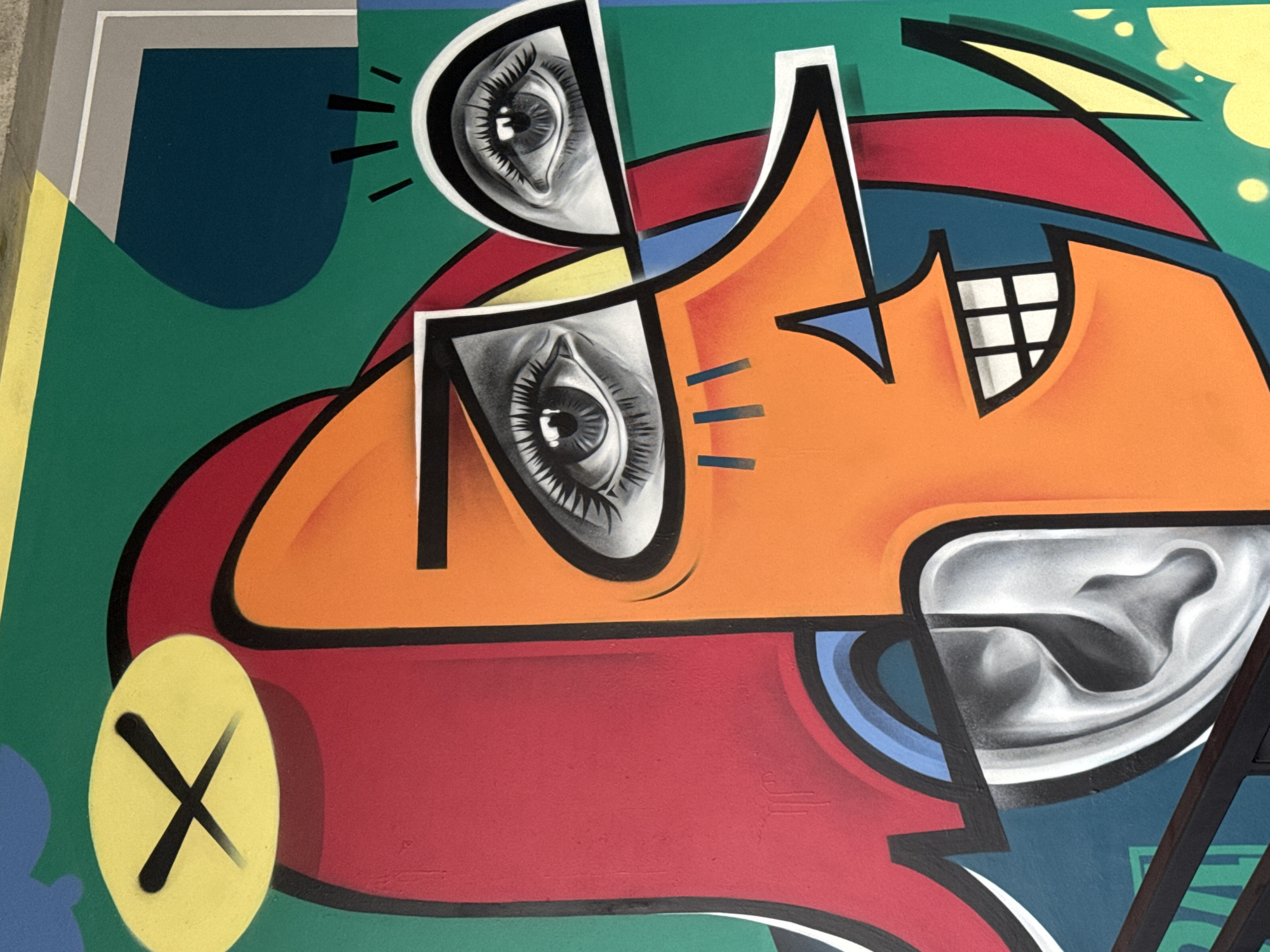 The image shows a colorful abstract mural. The artwork features exaggerated facial features, including two large, realistic eyes in stylized shapes, an orange face, teeth represented by squares, and a silver-colored area possibly depicting a cheek or ear. The mural has a background of green, blue, and yellow, with additional shapes and lines in various colors, including red, black, and white. A yellow circle with a black "X" is in the lower left corner. The location is in Sochi, Russia.