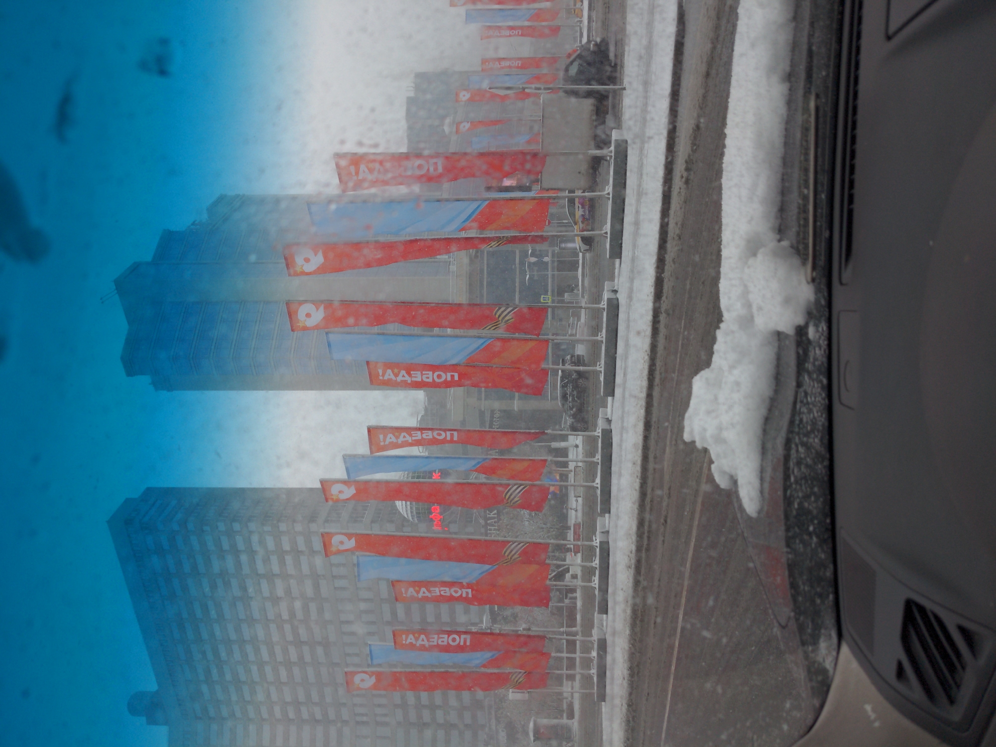 The image, taken from inside a vehicle through a rain-streaked windshield, captures an urban street scene in Unknown, Unknown. A row of flags, predominantly red and blue, line the street, affixed to metal poles. The text on the flags reads "!AA3EON" in a Cyrillic-like script. The background features high-rise buildings with a uniform grid of windows, suggesting a dense urban area. Snow accumulates along the roadside and the vehicle's exterior, visible on the side of the vehicle. The overall scene indicates a possible urban area during winter.