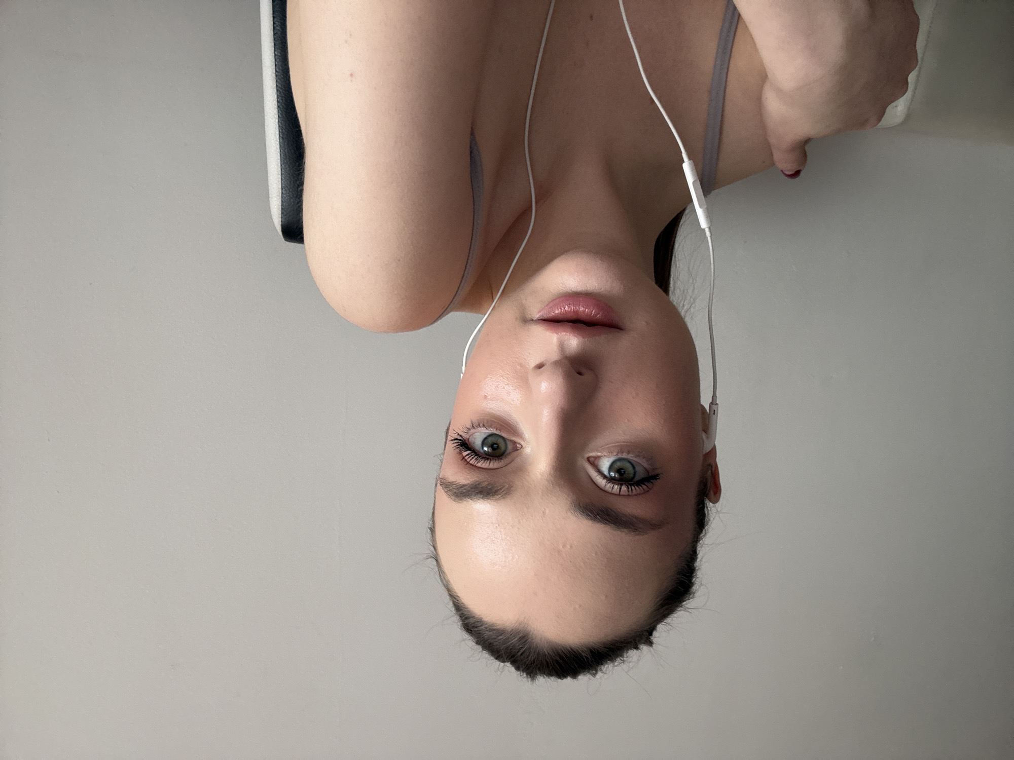 The image features a young woman in an indoor setting, captured from a low-angle perspective looking upwards towards the camera. Her head and upper torso are visible against a plain, light grey background. She has striking light-colored eyes, likely green or hazel, and well-defined eyebrows, with visible mascara on her eyelashes. Her dark hair is pulled back from her face, and her lips are slightly parted, showing a natural or lightly tinted color.
She is wearing white wired earphones, with the wires running down her chest and a small control unit visible near her collarbone. A light-colored strap, consistent with a top or sports bra, is visible on her shoulder. To the viewer's left, behind her right shoulder, part of a dark object with a white or light grey trim, possibly a chair back or headrest, is visible. The lighting is soft and even, indicating an indoor environment with good illumination. Her expression is neutral and direct, suggesting she is posing for the photograph. There is no visible text in the image. The scene is set in Kragujevac, Serbia.