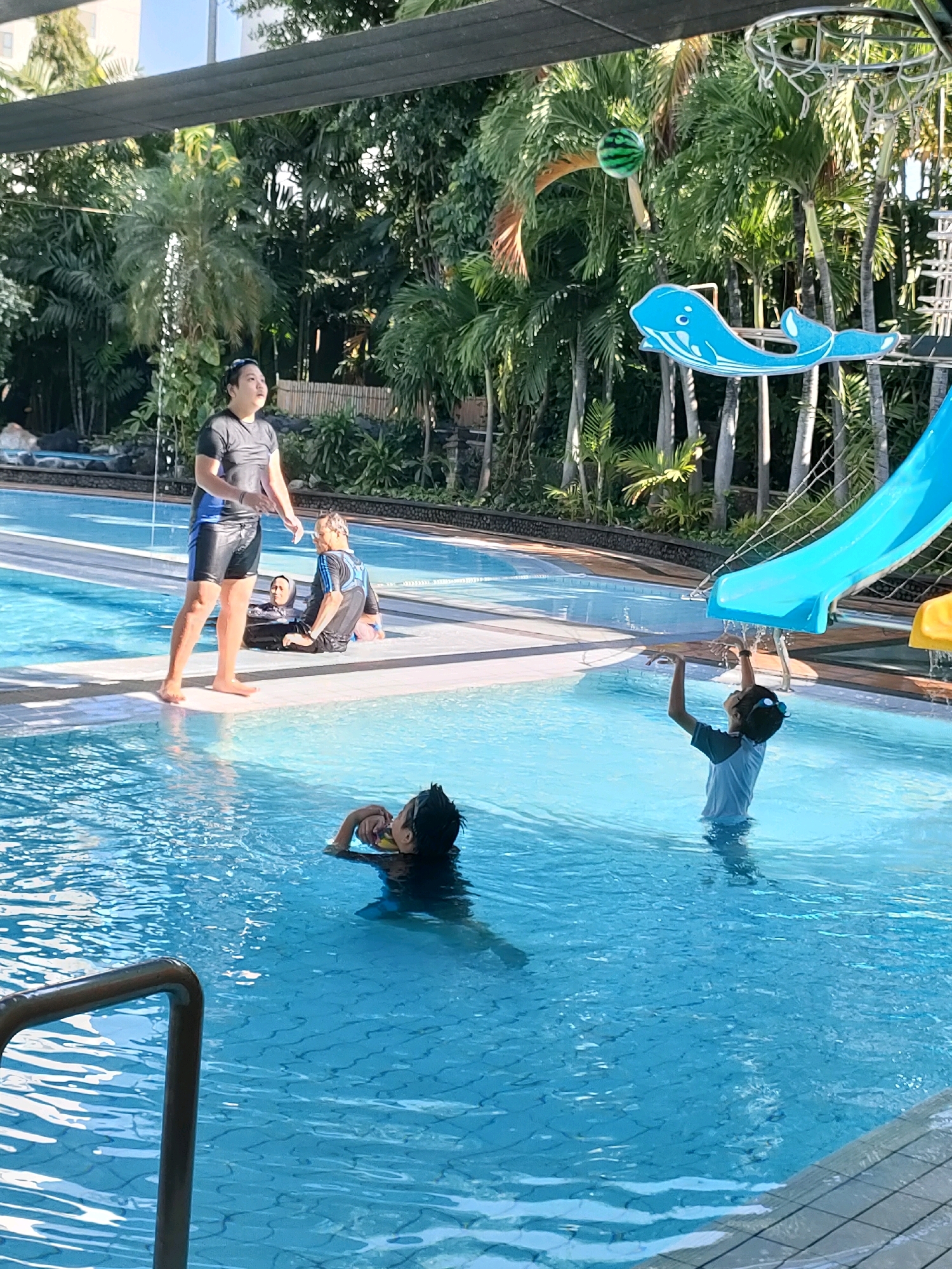 In Surabaya, Indonesia, at a swimming pool, a group of individuals is engaged in recreational activities. A person in the foreground, partially submerged in the water, holds a yellow object. To their right, a child in a light blue t-shirt and goggles stands in the water, reaching upwards. In the background, a person in a black and blue swimsuit stands at the edge of the pool, looking upwards. To their left, two individuals are seated in the water, one wearing a black and blue swimsuit and the other a dark garment with a head covering. The environment features palm trees, a blue water slide, and a basketball hoop with a striped ball in the air.