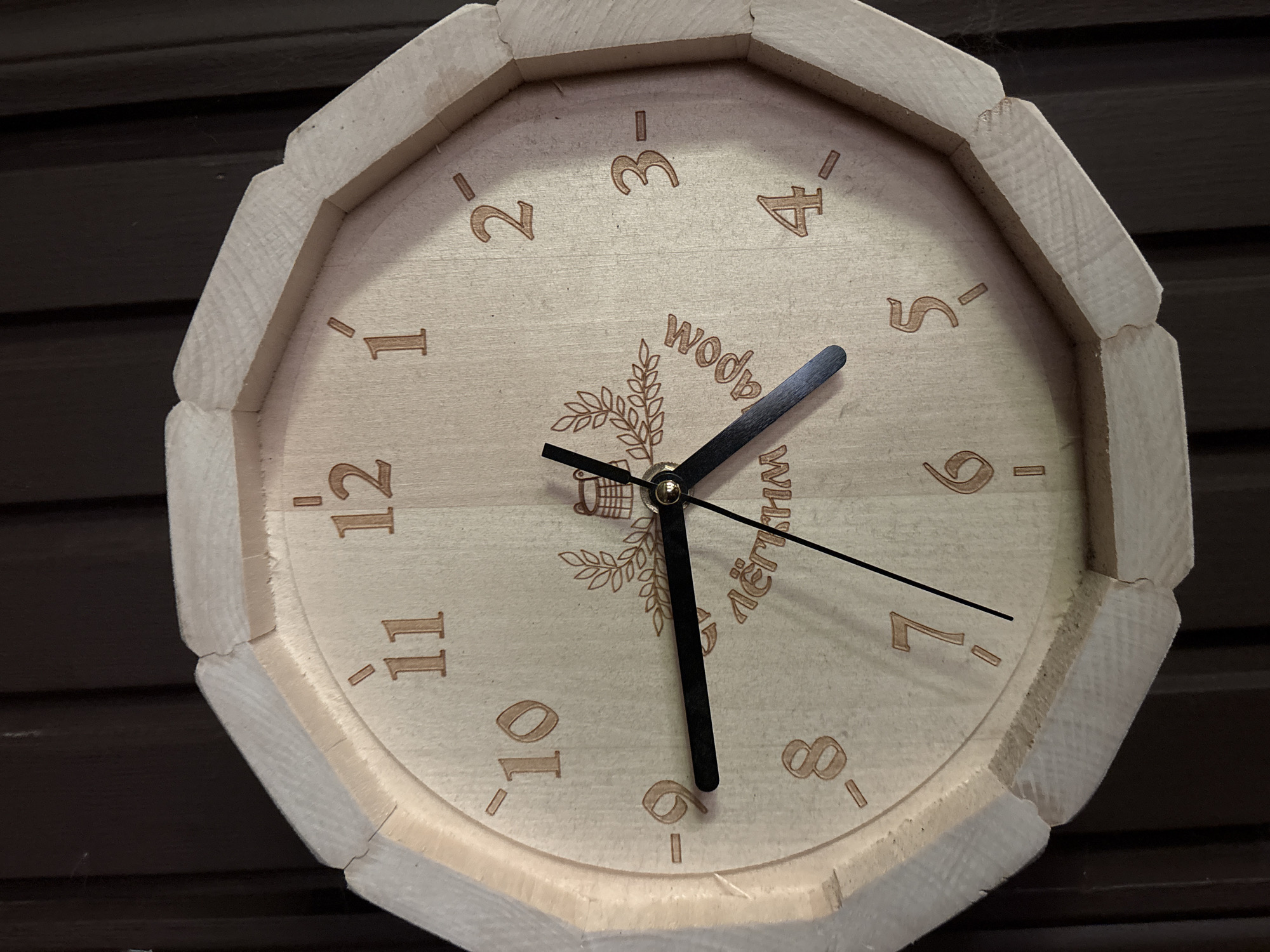 A wooden clock with carved numbers is hanging on a dark wooden wall. The clock face is light wood with darker brown numbers. The hands of the clock are black. The numbers on the clock are 1, 2, 3, 4, 5, 6, 7, 8, 9, 10, 11, and 12. There is also some text and a design carved into the clock face, which appears to be a laurel wreath and a basket. The clock has an octagonal frame made of lighter wood. The wall behind the clock has a vertical wood grain pattern. There are no people or animals visible in the image. The lighting is moderate, casting some shadows on the clock and wall. The clock appears to be in good condition.
This image was taken in Mikheevo, Russia.