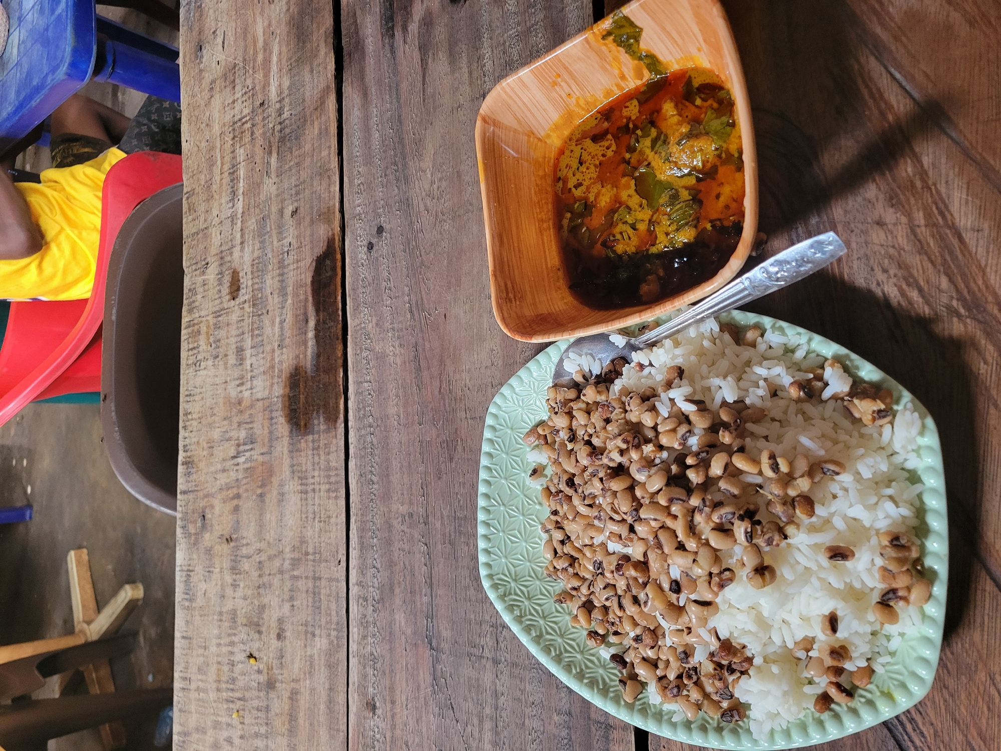 This image presents a top-down view of a meal served on a rustic wooden table, likely in Onitsha, Nigeria. The central focus is a light green, square-shaped plate with a raised, intricate pattern along its edge, filled with white rice and generously topped with cooked black-eyed peas (cowpeas). A silver spoon rests on the edge of the plate, partially obscured by the food.
Adjacent to the plate, a square-shaped wooden bowl contains a rich, reddish-orange stew or soup. This stew features visible pieces of green leafy vegetables or herbs, and a darker, possibly oil-based, liquid pools at the bottom.
The table itself is constructed from broad, natural wooden planks, showing prominent grain patterns, visible scratches, and some darker stains, suggesting it is well-used. In the background to the left, parts of a person are visible, wearing a bright yellow t-shirt and dark, patterned shorts, seated on a red plastic chair. A blue plastic chair and what appears to be a brown container are also partially visible, contributing to the casual and utilitarian setting. The natural lighting suggests the scene takes place during the daytime. There is no visible text in the image.